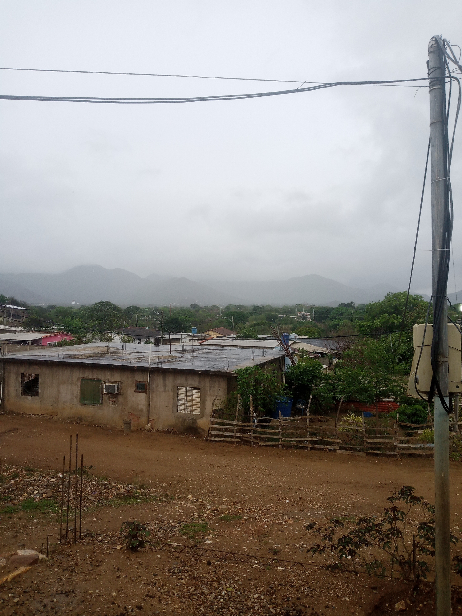 The image depicts a cloudy, overcast day in a residential area of La Tagua, Venezuela. In the foreground, a rough, unpaved dirt road with scattered rocks and debris leads towards a simple, concrete building with a flat, weathered roof. The building has several windows, some with bars and others with shutters. To the right of the building, a wooden fence separates the property from a yard with some sparse vegetation.
In the mid-ground, a cluster of houses with various roofing materials, including some with pink and red roofs, are visible. Green trees and foliage are abundant, suggesting a humid climate. Further in the background, a range of low, mist-shrouded mountains forms the horizon.
A utility pole with numerous wires and a light-colored electrical box stands prominently on the right side of the image, near the foreground. The overall impression is one of a somewhat rural or underdeveloped urban setting under a gray, potentially rainy sky. No people are visible in the scene.