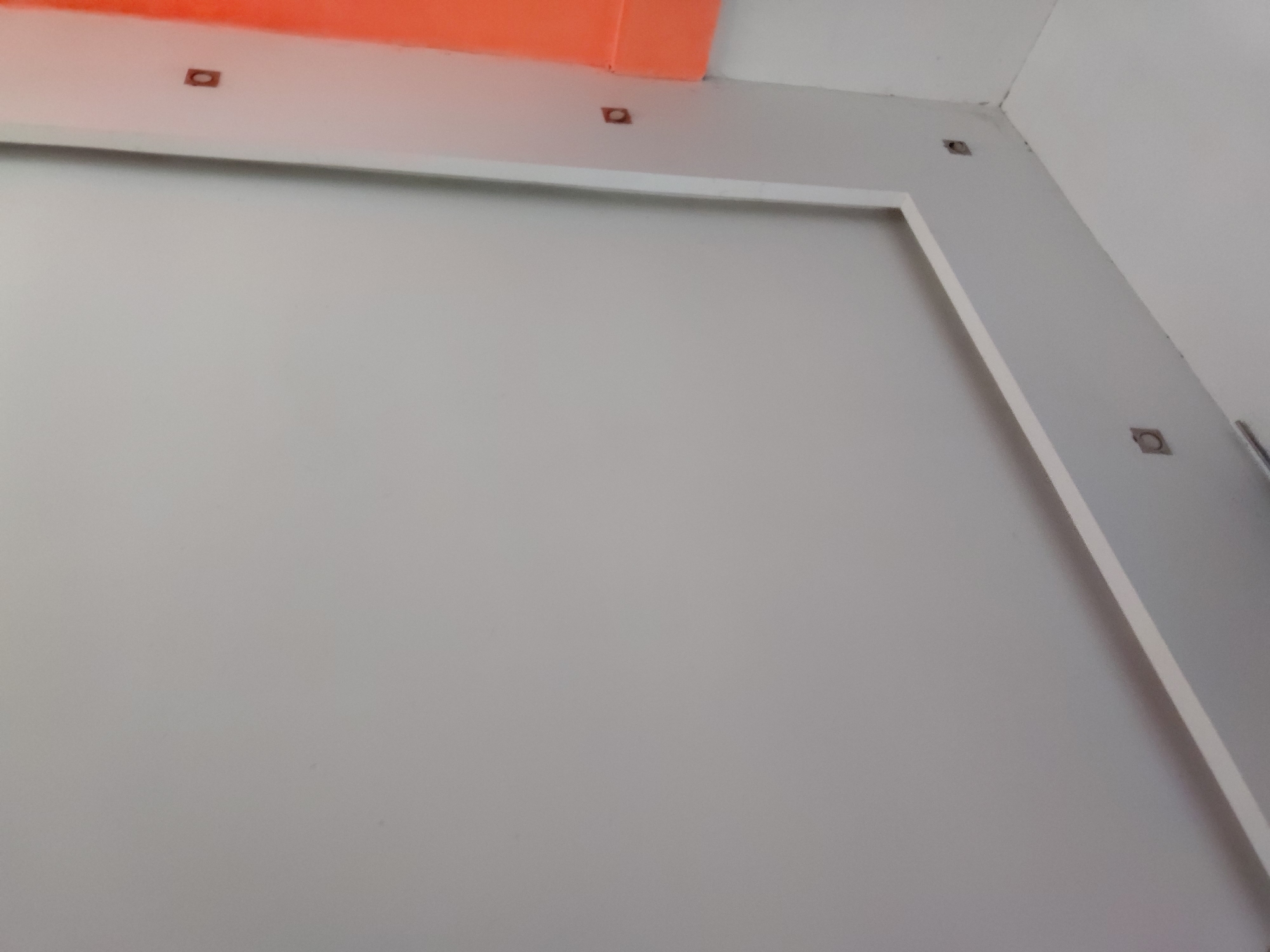 The image captures an interior architectural detail, specifically a corner where a light grey wall meets a white ceiling, featuring a striking bright orange section. A white rectangular trim or molding frames a large, plain section of the light grey wall. Several square-shaped cutouts, which appear to be incomplete electrical boxes or recessed fixture housings, are visible on the surfaces. Three such cutouts are positioned along the top edge of the grey wall, near the orange section and the ceiling. Another two are seen further along the grey wall and in the corner, suggesting preparations for lighting or other installations. In the bottom right corner, a glimpse of a vertical, metallic object, possibly a pipe or railing, can be seen. The space appears to be under construction, recently renovated, or a newly built structure awaiting final fixtures. The lighting is even, suggesting an indoor setting, possibly during the day. There are no people visible, nor any discernible text. The scene is clean and orderly, reflecting a modern or minimalist design aesthetic. The location context provided is Santa Ana, Venezuela.