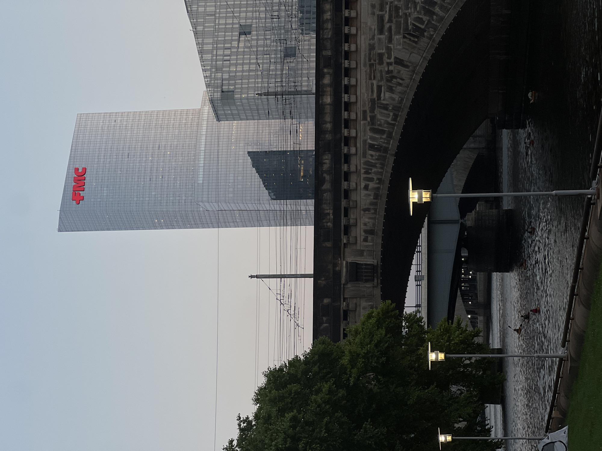 The image depicts an urban waterfront scene in Philadelphia, Pennsylvania, USA, likely in the evening or early morning due to the soft lighting and clear sky. Dominating the background are two modern glass-clad skyscrapers. The taller of the two, on the left, features a prominent red "FMC" logo at its apex. Adjacent to it, a shorter, wider glass building is visible.
In the mid-ground, a large, arched stone bridge spans a body of water, likely the Schuylkill River. Beneath this bridge, another, more modern bridge structure is visible, with its supporting pillars and walkway illuminated. Overhead, a complex network of train tracks and overhead power lines crisscrosses the scene.
In the foreground, the bank of the river is lined with green grass and a paved walkway. Two modern street lamps with rectangular lighting elements stand on the walkway, illuminating the area. In the water, several people are engaged in kayaking, with two individuals visible in kayaks near the center of the frame, and others further in the distance. A small, white tent-like structure is visible on the grassy bank to the left. The water is rippled, reflecting the overcast sky and the structures above. The overall atmosphere is one of urban tranquility, with human activity integrated into the cityscape.