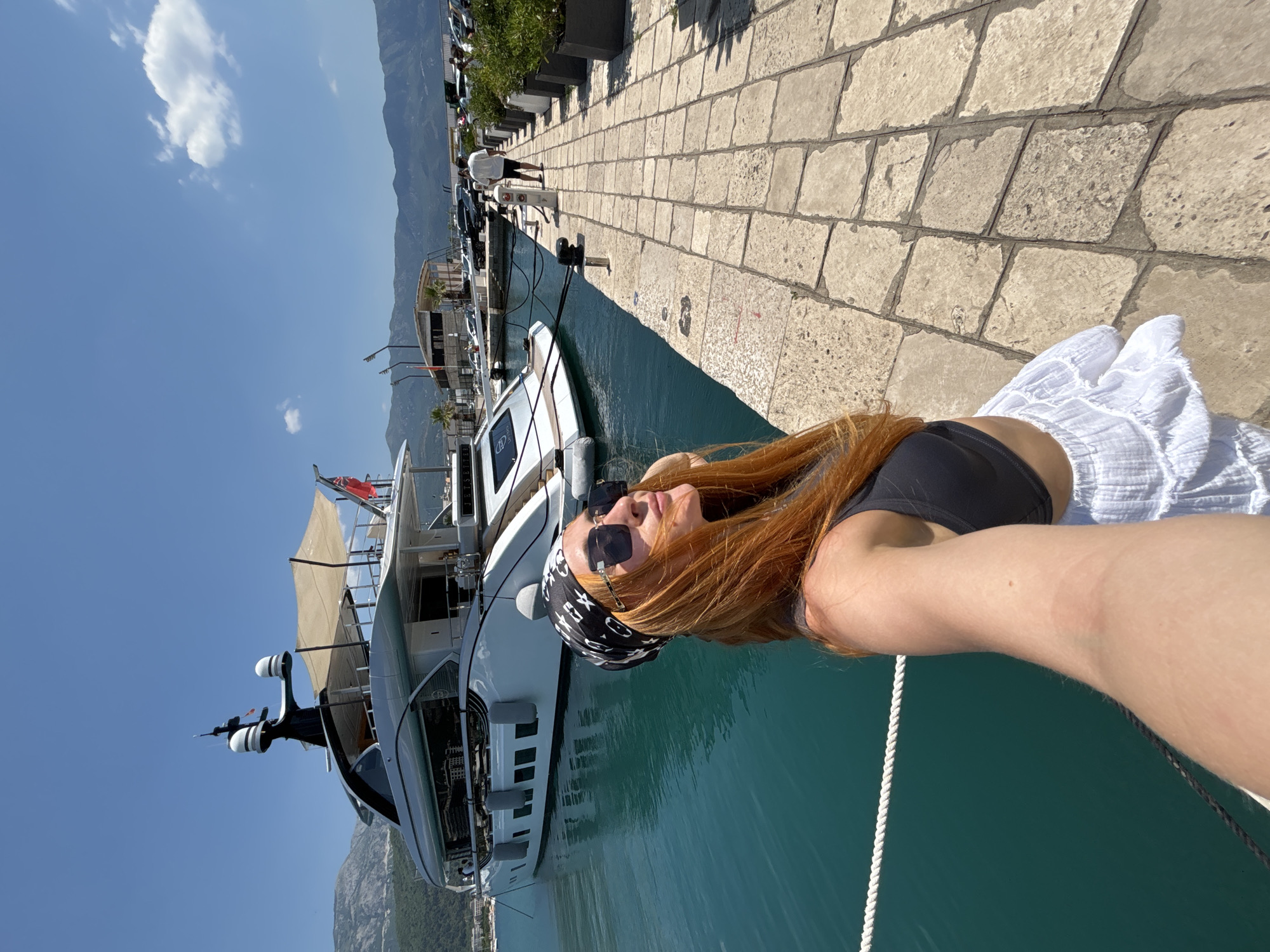 A woman with long auburn hair is taking a selfie by a marina on a sunny day. She is wearing a black bikini top and a white skirt. She has sunglasses on and a black and white bandana on her head. She is leaning on the railing of a large white yacht. The yacht is docked next to a stone walkway. In the background, there are more boats, mountains, and a clear blue sky. The scene appears to be in Budva, Montenegro, as indicated by the provided location context. The lighting suggests it is daytime, and the clear sky indicates fair weather. The woman's pose and smile suggest a relaxed and happy mood. There is no visible text in the image.