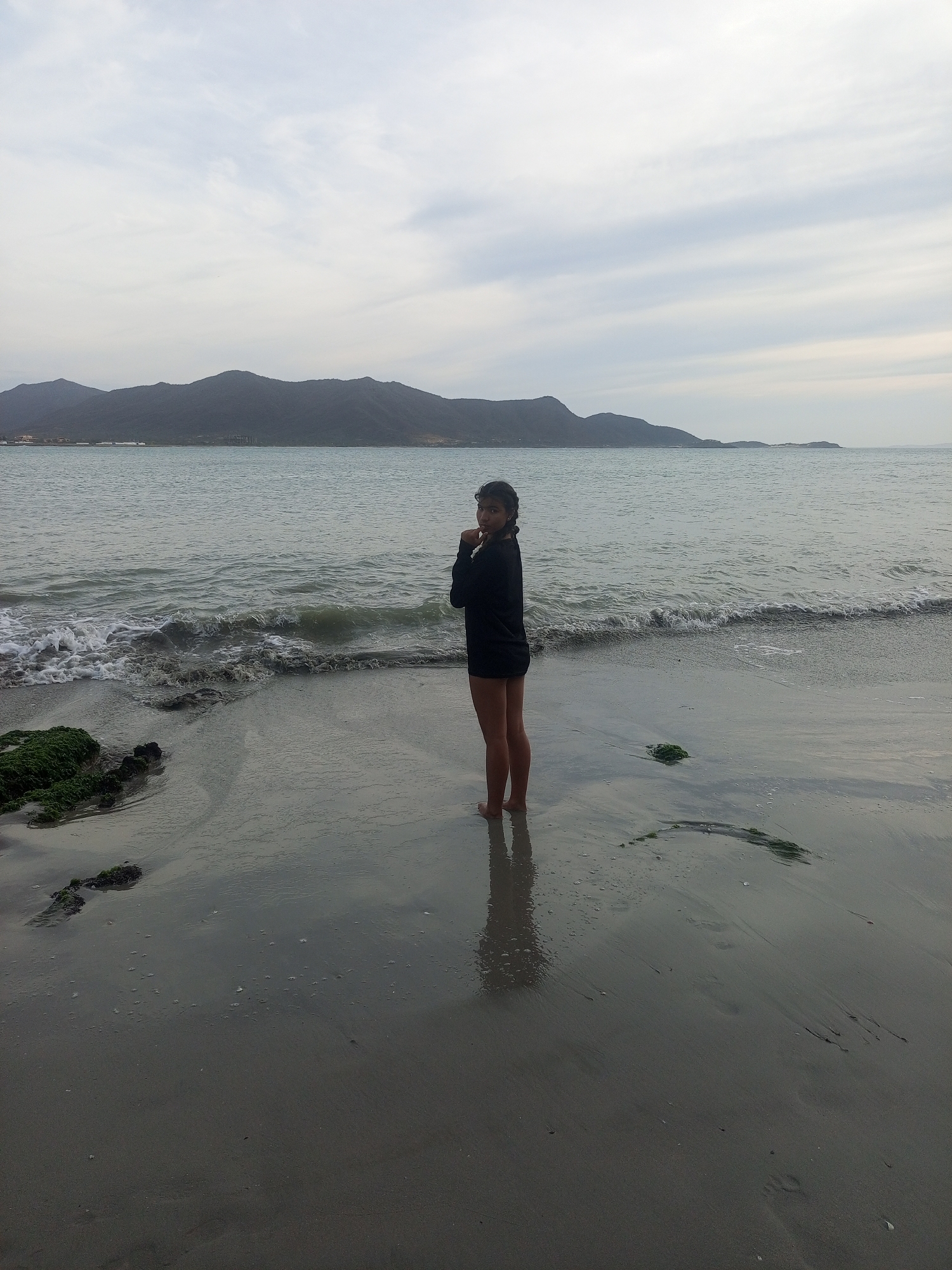 A young person stands on a sandy beach in Juan Griego, Venezuela. The individual is wearing a dark long-sleeved garment and dark shorts, facing away from the sea. Behind the person is the ocean with gentle waves. The horizon shows hills or mountains under an overcast sky. Patches of green algae or seaweed are scattered on the wet sand. The scene conveys a calm, somewhat cool atmosphere.