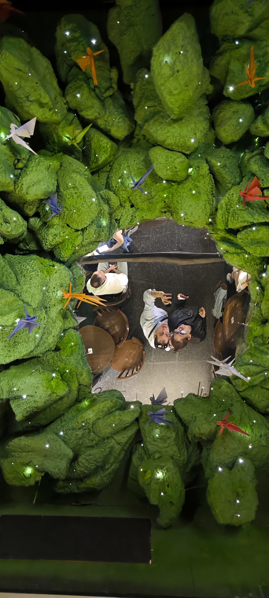 Captured from a high-angle perspective, the image reveals an interior space reminiscent of a constructed diorama. The scene is framed by rough, textured green forms, possibly artificial rocks or moss, adorned with small, bright lights that suggest a magical or fantasy setting. Multicolored origami birds are suspended amidst the green textures, adding to the whimsical feel.
Several people are present in the center of the frame. One individual is seated on a simple wooden chair, facing away from the camera. Closer to the foreground, two individuals are standing and appear to be posing for a photograph. One is wearing a light-colored shirt or jacket, while the other is dressed in black. They are positioned close together, suggesting a friendly or intimate relationship. A few other figures are barely visible at the edge of the frame. There are also wooden tables and chairs, set to the side of the scene.
Based on distinctive elements of decor and artistic features, it can be deduced that the location of the image is inside a restaurant called Ikibana Parallel in Barcelona, Spain. Ikibana restaurants are known for their distinctive interiors, which often incorporate nature-inspired elements and whimsical decorations.