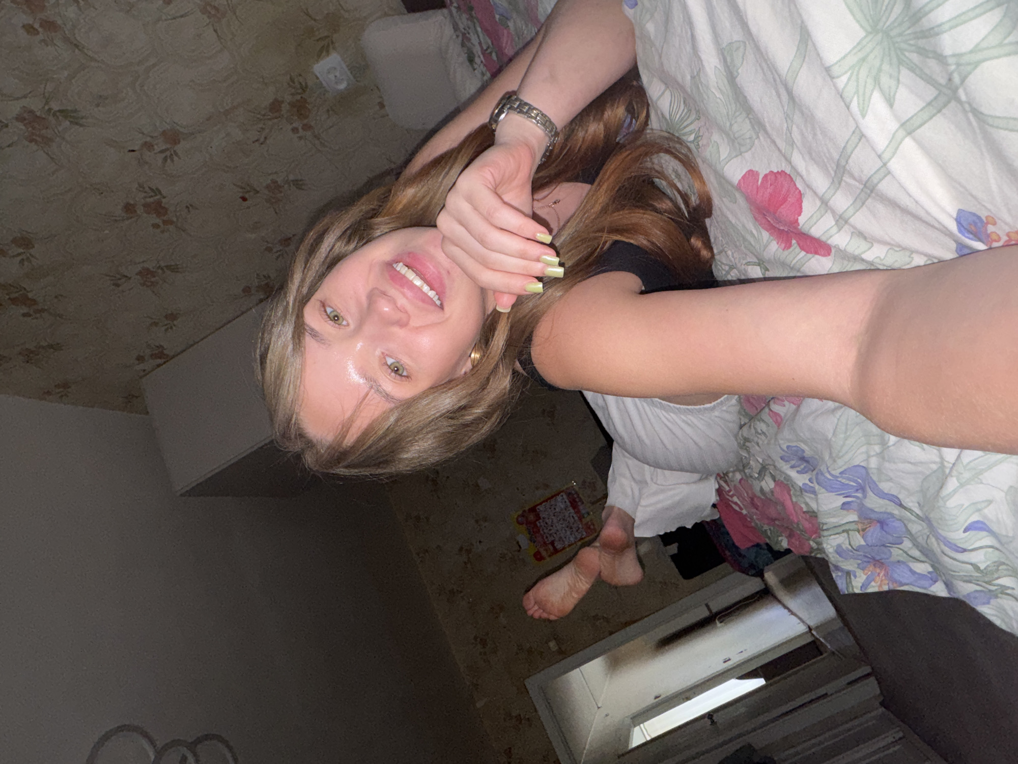 The image depicts a young woman lying on a bed in a room. She is facing the camera and smiling, with her right arm extended to take the photo. She has long, wavy brown hair and green eyes. She is wearing a black top and white pants. Her fingernails are painted a light green color. On her wrist, she is wearing a silver watch. Her feet are visible, resting on the bed. The bed is covered with a floral patterned comforter. The room appears to be dimly lit. In the background, there is wallpaper with a floral pattern, a doorway, and a closet filled with clothes. The location is identified as Chaikovsky, Russia.