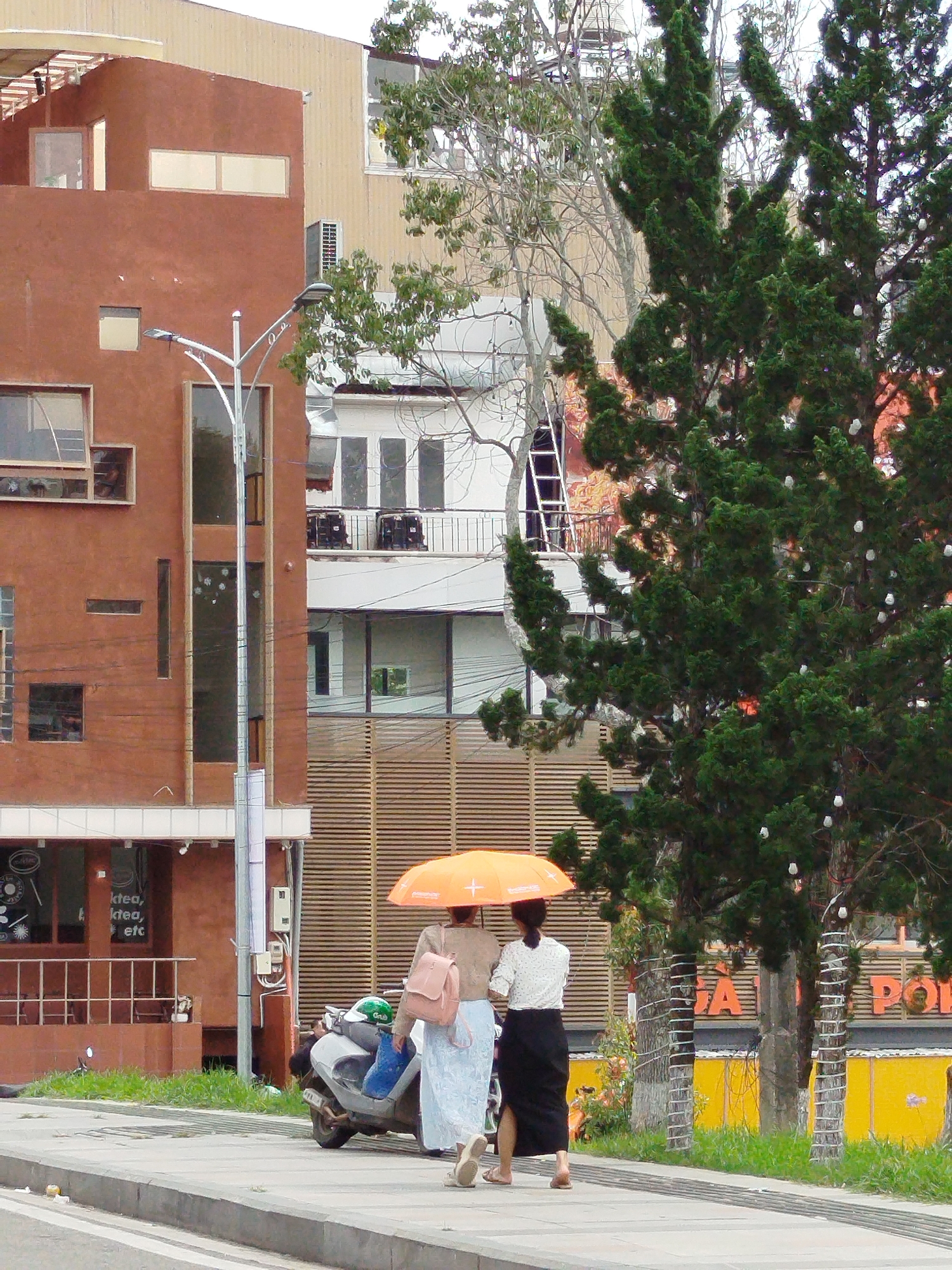 The image depicts a street scene in Phường Xuân Hương, Đà Lạt, Vietnam, focusing on two women walking with an umbrella. The woman on the left, facing away from the camera, is wearing a beige jacket and a light blue skirt, carrying a pink backpack. The woman on the right, also facing away, wears a white patterned blouse and a black skirt. They are walking on a sidewalk next to a road, with a motorbike parked on the left side of the sidewalk. In the background, there are multi-story buildings with distinct architectural features, including brown and beige facades, balconies, and windows. Lush green trees, one particularly prominent evergreen tree with white lights, are situated between the buildings and the sidewalk. The overall environment suggests a bright day, with the orange umbrella providing a splash of color against the more subdued tones of the buildings and vegetation. The presence of advertising on the buildings and the general urban setting indicate a commercial or residential area.