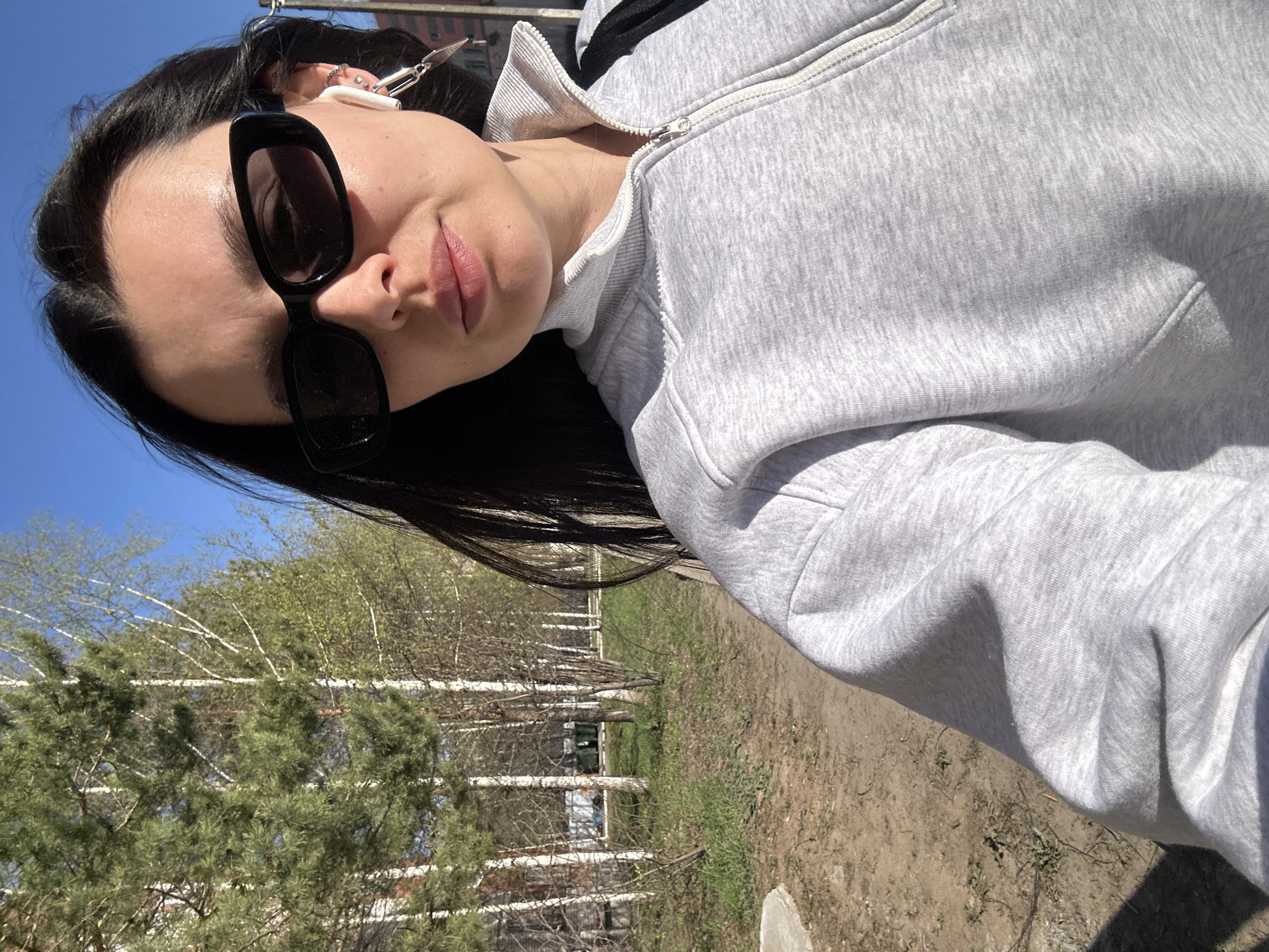 A woman wearing black sunglasses and a light grey zip-up sweatshirt is taking a selfie on a path in a park. The park appears to be in Chaikovsky, Russia, considering the trees and vegetation visible in the background. The ground is a mix of dirt and grass. There are trees lining the path, some with bare branches and others with green foliage. A building is visible in the distance behind the trees. The sky is blue.