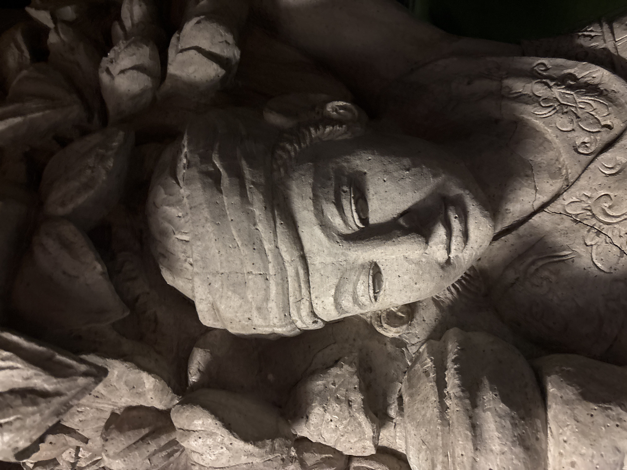A close-up shot depicts a weathered stone carving, possibly a relief or sculpture, featuring a human figure. The figure is lying on their back, head tilted to the left, with visible facial features including eyes, nose, and mouth. Details around the figure suggest foliage or cloth and decorative patterns. The photo was taken in Kuta Selatan, Indonesia. The image appears to be of an ancient or historical artifact due to the style of carving and state of disrepair. The figure's clothing and the overall aesthetic suggest it might be of religious or cultural significance.