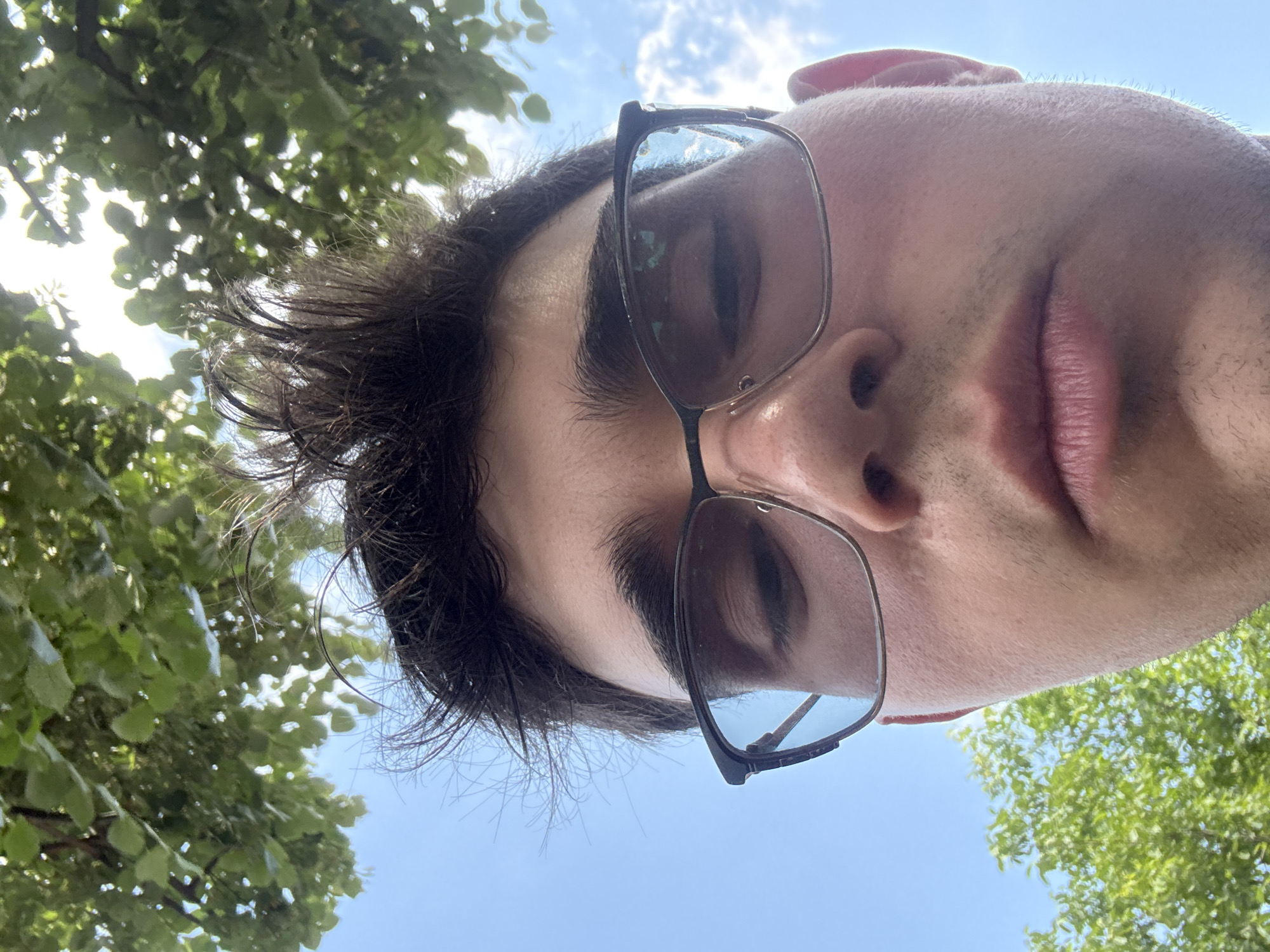 The image is a low-angle, close-up selfie of a young Caucasian male with dark, messy hair and light skin. He is wearing dark-rimmed glasses. His mouth is slightly open, and he is looking downwards. The background consists of bright green foliage from trees against a clear blue sky with some scattered white clouds. The lighting suggests it is daytime, possibly late morning or early afternoon. There are no identifiable landmarks or context to place this image within the city of Isperih, Bulgaria, or any other specific location. The scene appears to be outdoors, likely in a park or natural setting.