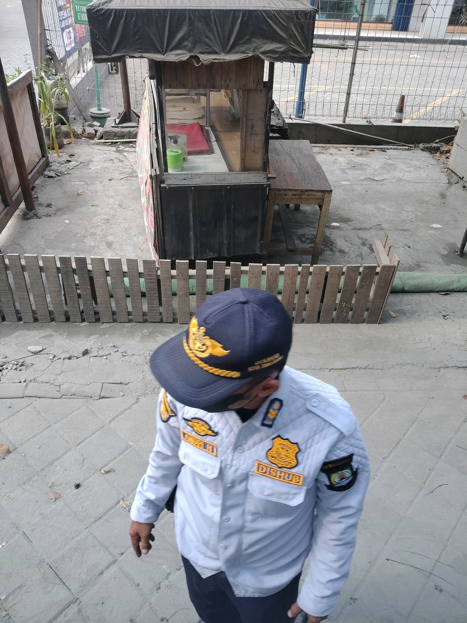 The image shows an outdoor scene in Tangerang, Indonesia, with a focus on a person wearing a light blue uniform and a dark blue cap. The person appears to be a security guard or public official, indicated by the patches and emblem on their uniform, including one that reads "DISHUB" (likely referring to the transportation agency). The guard is facing away from the camera, walking on a paved walkway. In the background, there is a small wooden food stall with a dark canopy and glass display, accompanied by a wooden bench. A low wooden fence separates the walkway from a dirt and debris-covered area. Beyond the fence, there is a fenced-off lot with some construction materials and a traffic cone. The overall environment suggests a public area in a city, possibly near a market or roadside.