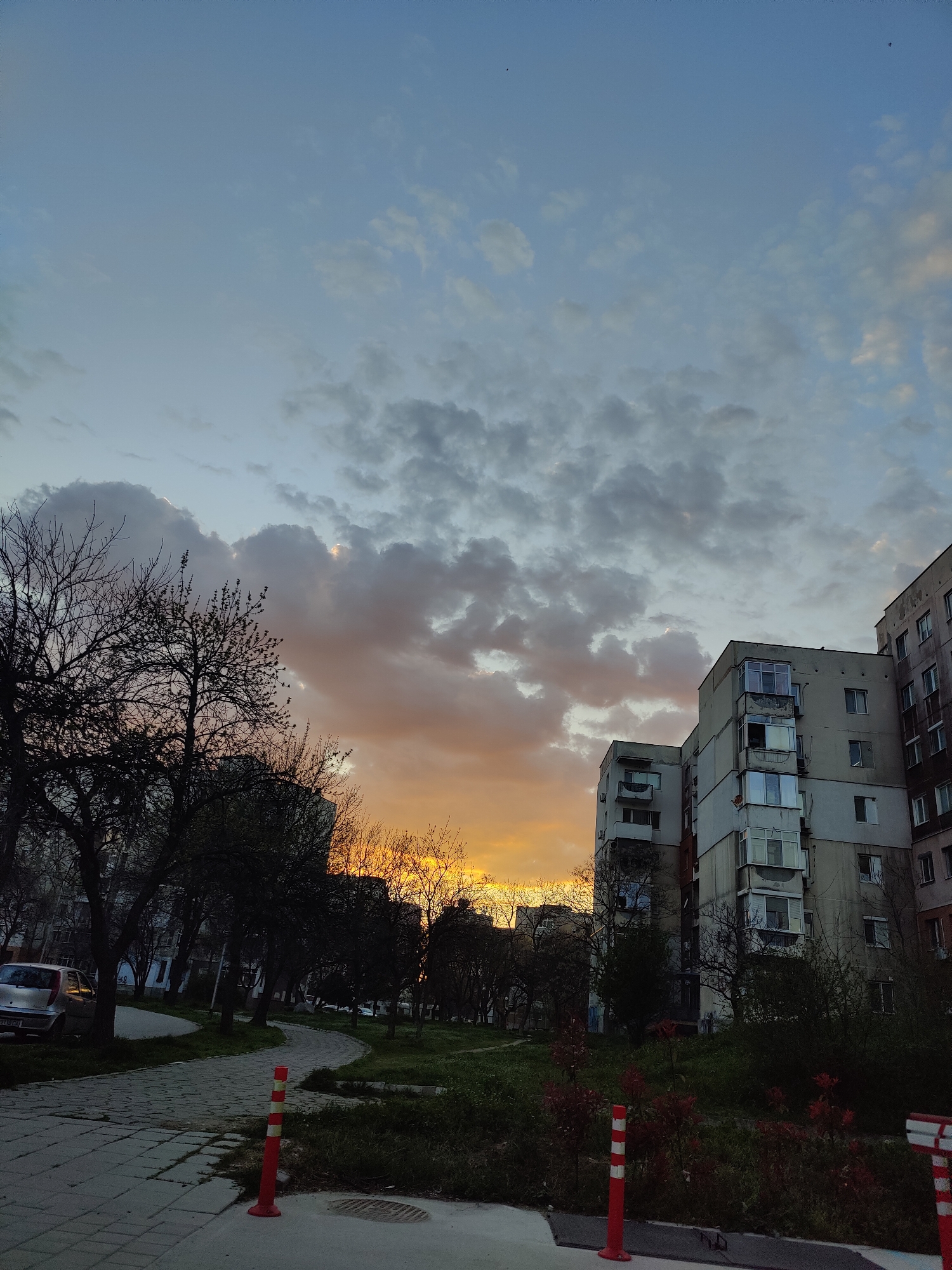 The image shows an urban scene in Plovdiv, Bulgaria, at sunset. The sky transitions from blue at the top to orange near the horizon, with grey clouds dominating the central area. Bare trees line the left side of a paved pathway, where a silver car is parked. On the right, several concrete apartment buildings rise, showing signs of age and disrepair. A grassy area with some shrubbery separates the pathway from the buildings. Orange traffic pylons are sparsely placed along the edge of the path. No people are visible.