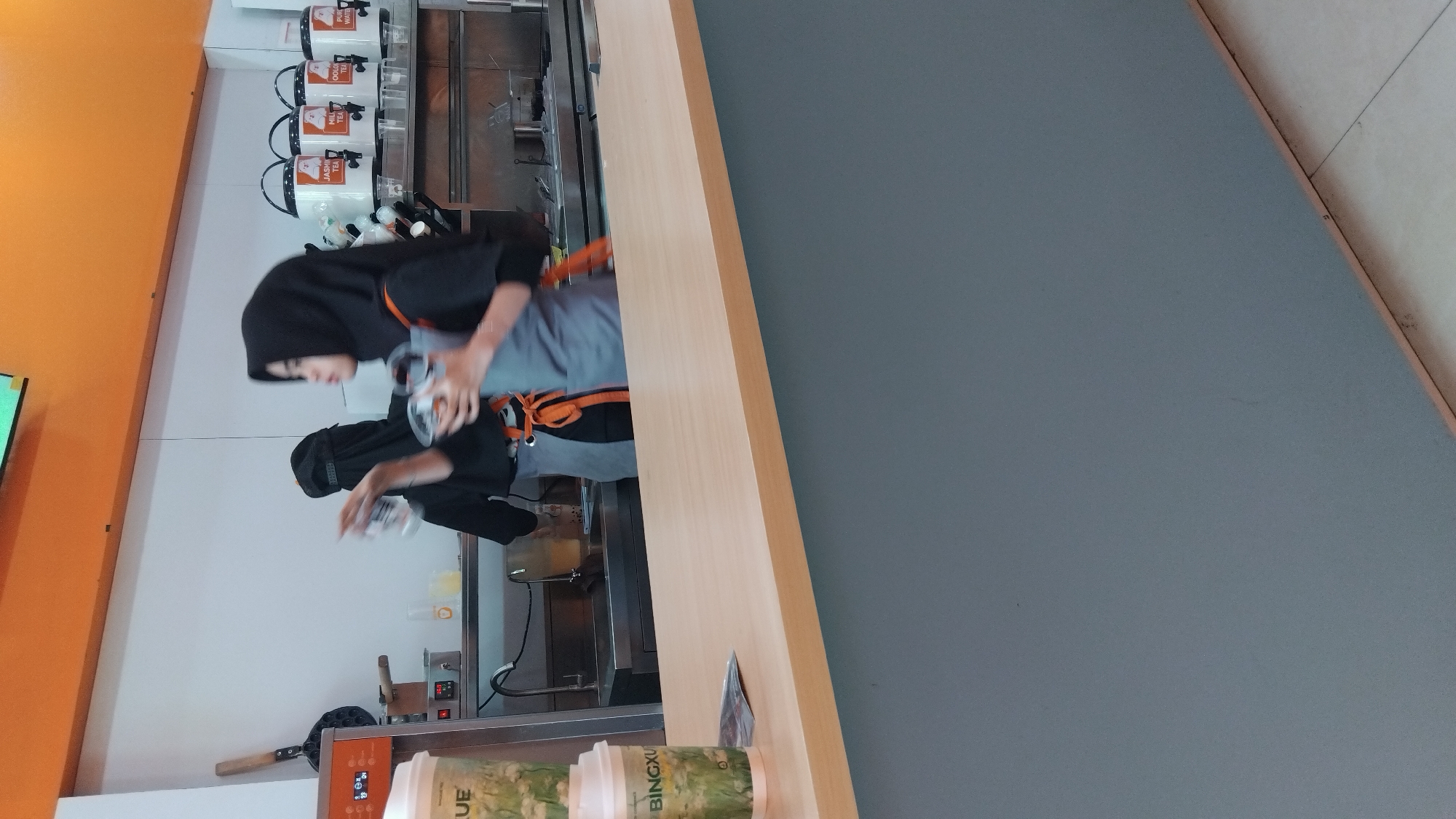 In the city of Batang, Indonesia, a scene unfolds at what appears to be a beverage or food service counter. The foreground is dominated by a light wood-colored counter. Behind the counter, two individuals are visible, both wearing black tops and headscarves. One person, positioned further back and to the left, is partially obscured but appears to be interacting with equipment on the counter, possibly a waffle maker. The other person, closer to the center and right, is wearing a gray apron over their black top and is holding a clear plastic cup with a lid. They seem to be preparing or serving a drink.
Behind the workers, the environment features a stainless-steel countertop with a sink on the left. Various containers, including what look like large beverage dispensers with lids labeled "JASMINE TEA," "MILK TEA," "OOLONG TEA," and "PURE WATER," are arranged on a shelf above the countertop. Several stacked clear plastic cups are also visible. A light-colored wall forms the backdrop, with an orange band across the top. Two takeaway cups, one with the label "UE" and another labeled "BINGXU," are placed on the counter in the foreground. The lighting suggests an indoor commercial setting.