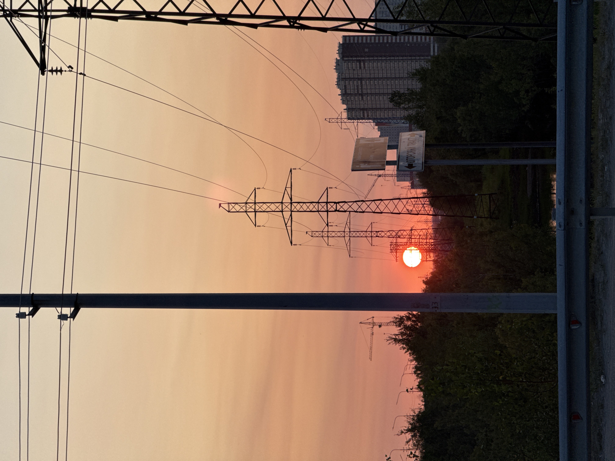 The image depicts a sunset over a suburban area of Surgut, Russia, characterized by industrial and residential structures against a backdrop of warm-toned sky. In the foreground, a silver guardrail runs horizontally, partially obscuring a road or path. A tall, grey utility pole stands prominently on the left, with its shadow cast across the frame. To the right, a large, black lattice electricity pylon dominates the view, with multiple wires stretching across the sky.
Further into the scene, a series of electricity pylons are visible, receding into the distance. The sun, a bright orange orb, is partially obscured by one of these pylons and the surrounding trees, casting a vibrant glow. In the middle ground, dense green foliage forms a line, with a body of water visible beneath it on the left. A road sign with Russian text, indicating "Югорский ТРАКТ" (Yugorsky Tract), is mounted on a pole. Behind the trees and pylons, several tall, modern residential buildings with numerous windows are visible, suggesting an urban or peri-urban setting. A construction crane can be seen in the far left background. The overall impression is one of an industrial landscape at dusk, with the natural beauty of the sunset creating a striking contrast.