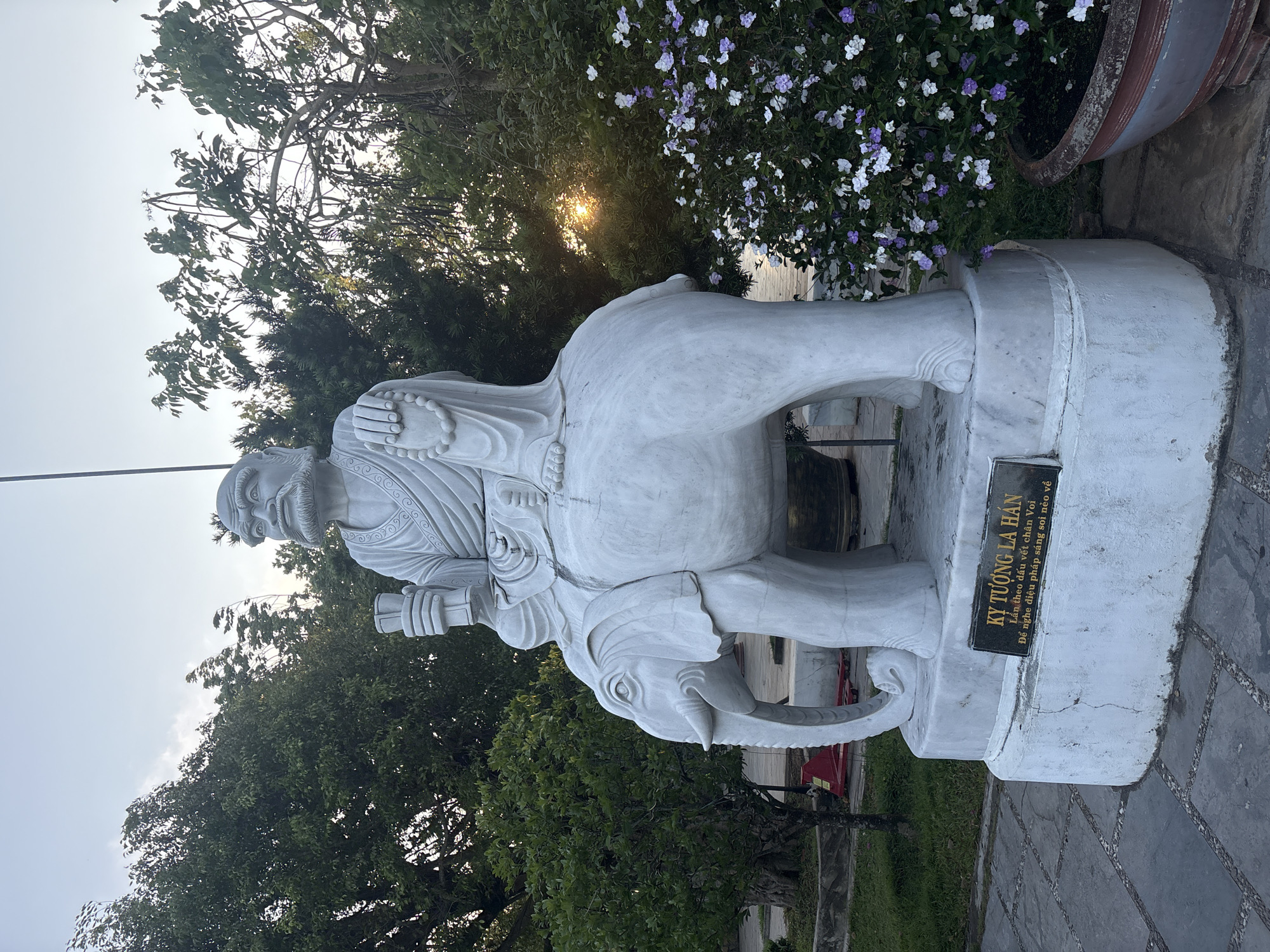 This is a daytime shot of a statue of an Arhat (La Hán) riding an elephant, located in Đà Nẵng, Vietnam. The white statue is positioned outside, surrounded by trees and flowers. A sign on the base of the statue reads "KỴ TƯỢNG LA HÁN" and a Vietnamese text. The statue is detailed, showing the Arhat with a beard and wearing traditional robes. The elephant features detailed tusks and ears. The statue is placed on a stone base. The ground is paved with stone tiles. In the background, sunlight filters through the trees.