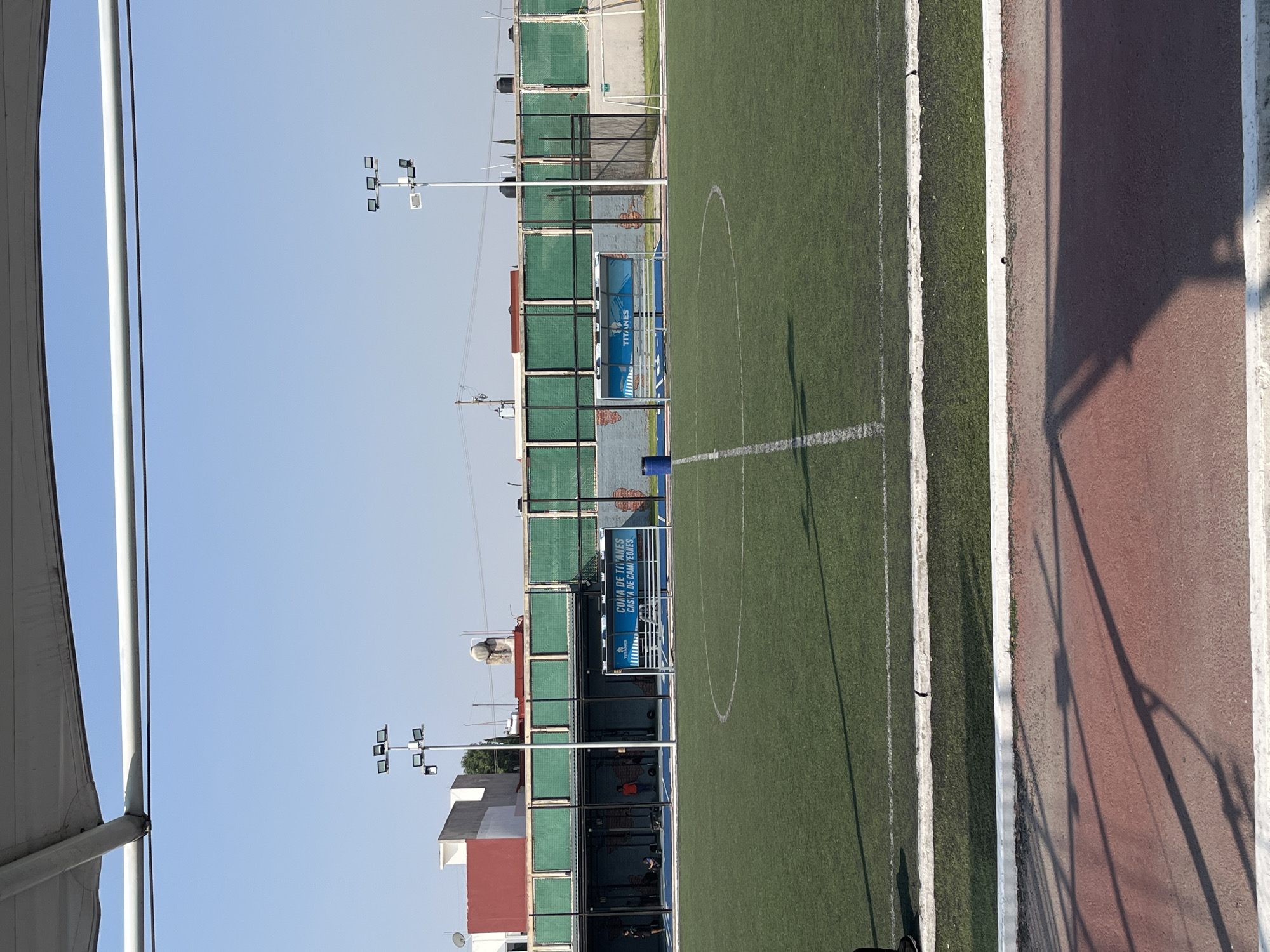 The image shows a sports field with artificial turf, a running track, and fencing around the perimeter. There are light poles with multiple lamps on each. Adjacent to the field is a building with multiple levels, covered in green mesh. A structure with a curved, white awning is visible in the foreground. The sky is clear and blue. The location appears to be an urban area.