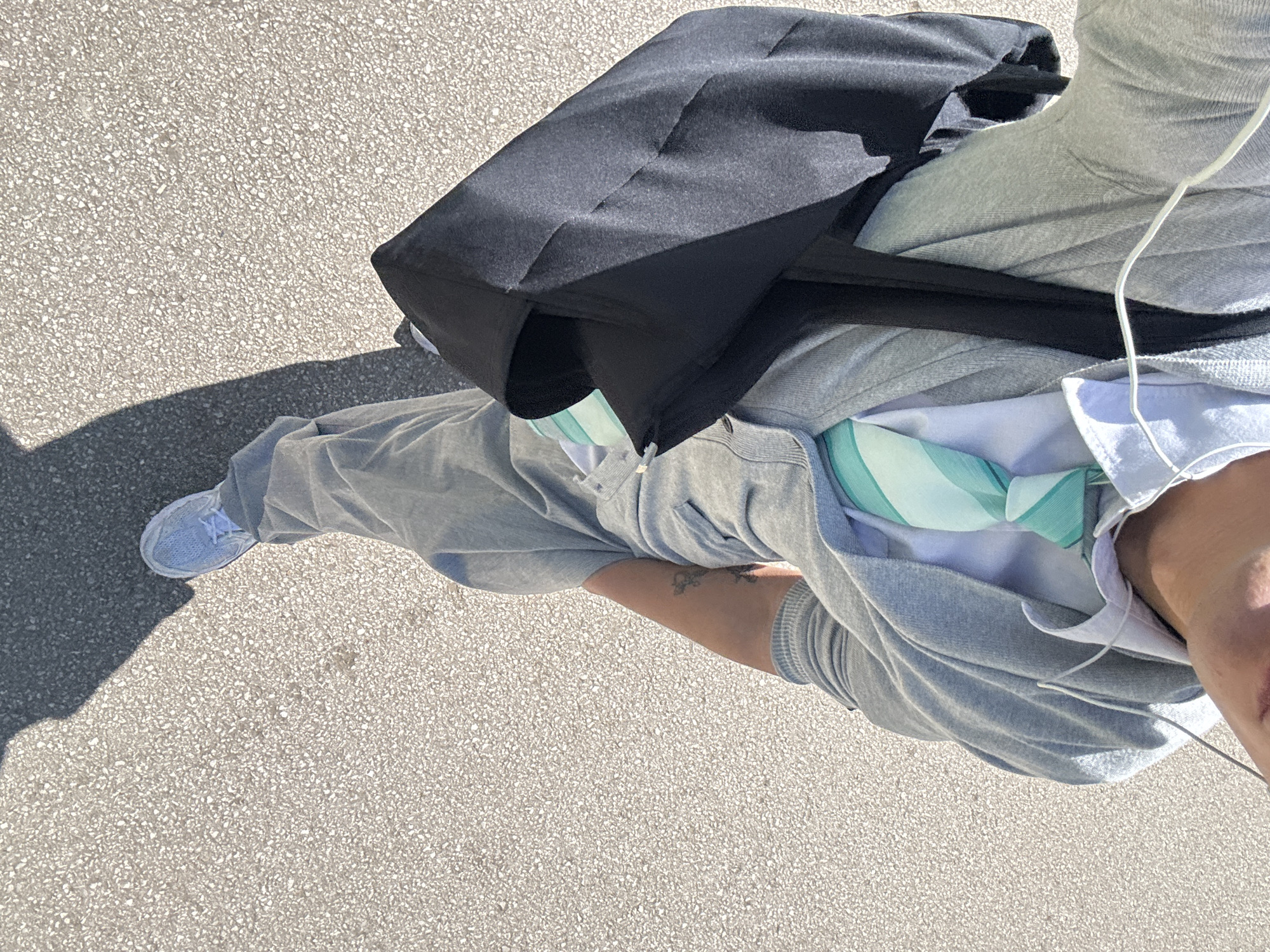 The image captures a top-down view of a person on a paved surface in bright daylight. The subject, likely female given the visible skin and tattoo, is primarily shown from the torso down, facing away from the camera. They are wearing a casual outfit consisting of a grey sweatsuit, including a hooded top and sweatpants. White sneakers are visible on their feet.
A black shoulder bag or backpack is carried across their back and shoulder. White wired earbuds are draped around their neck and chest, suggesting they are listening to audio. A light-colored garment, possibly a white t-shirt or undershirt, is visible beneath the grey hoodie. Around the neck or tucked into the top, a distinctive teal or mint green patterned fabric, possibly a scarf or a face mask, adds a pop of color to the otherwise neutral outfit. A small, dark-colored tattoo is visible on the inner forearm or bicep area of the person's right arm.
The ground is a light grey, textured asphalt or concrete pavement, characteristic of urban pathways. Strong, dark shadows are cast on the ground to the left of the person, indicating direct sunlight, likely mid-morning or afternoon. There are no other discernible objects or people in the frame. The scene is set in Sofia, Bulgaria. No visible text is present in the image.