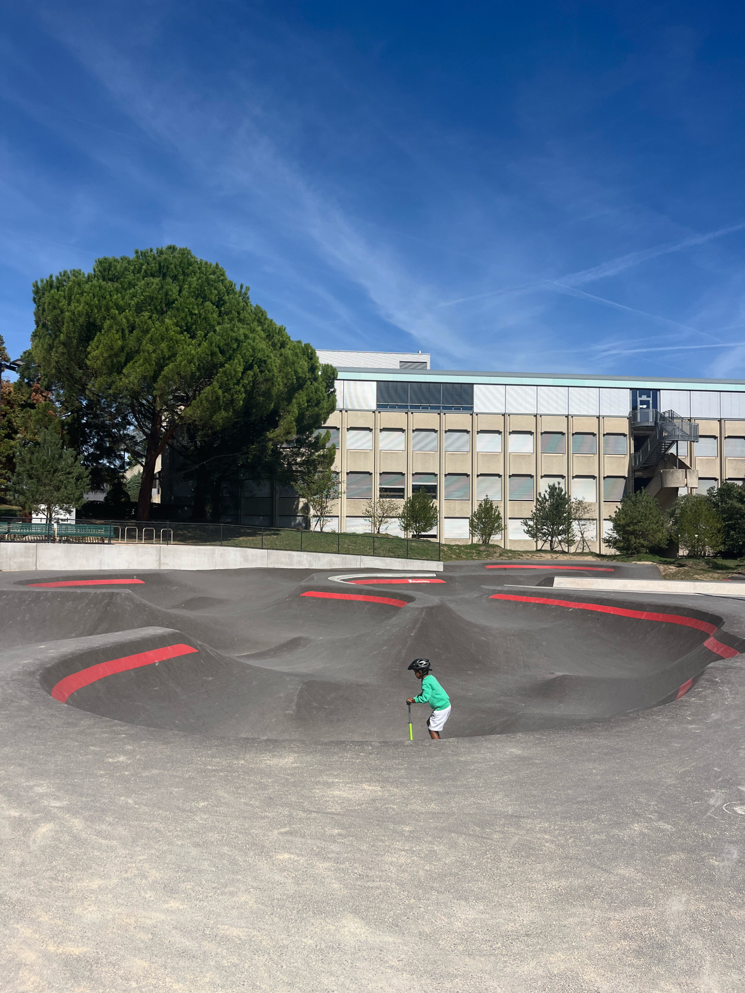 A young child wearing a green sweatshirt, white shorts, and a black helmet is riding a scooter on a black asphalt pump track with red accents. The pump track is located outdoors, in front of a building with multiple windows. Behind the building are trees, and the sky is blue with some wispy clouds. The scene appears to be in an urban or suburban area. Specifically, this image was taken at the Eaux-Vives pump track in Geneva, Switzerland.