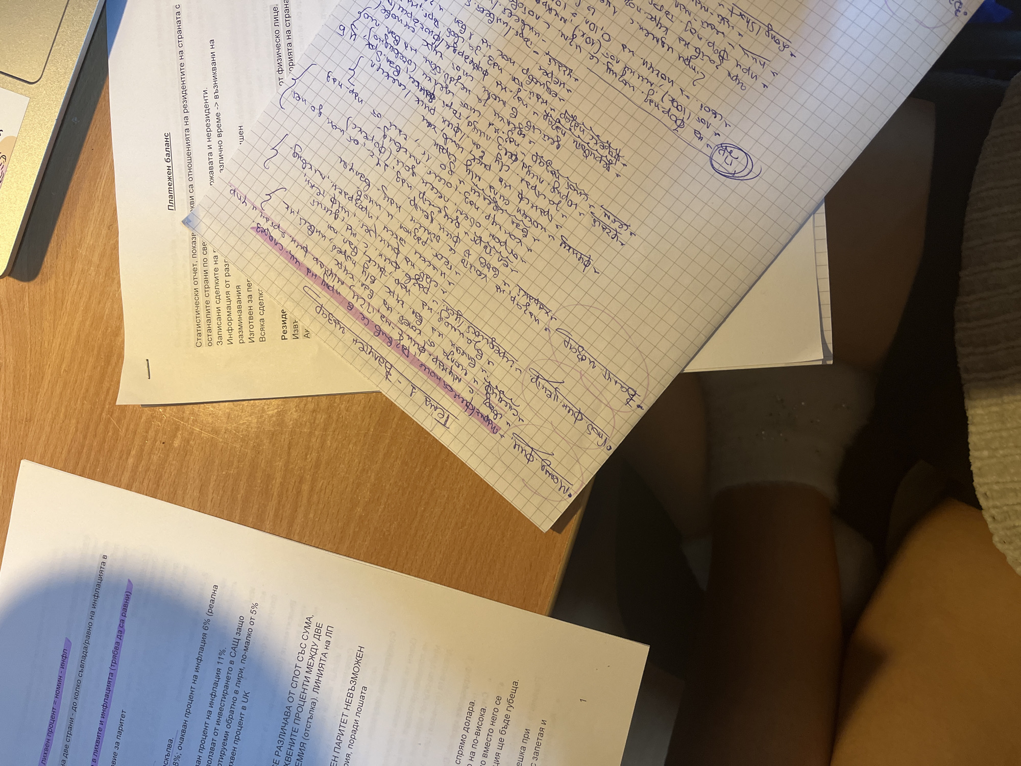 The image shows a desk with various papers and a laptop. On the left, there are documents in Bulgarian with economic and financial terms highlighted. A handwritten page with notes in Bulgarian is at the center, with headings such as "Валутен пазар" (Currency Market) and bullet points discussing financial concepts. The lower right corner reveals a glimpse of a person's legs and white socks. The overall scene suggests someone studying or working with financial materials. There is no indication of a location within Sofia, Bulgaria.