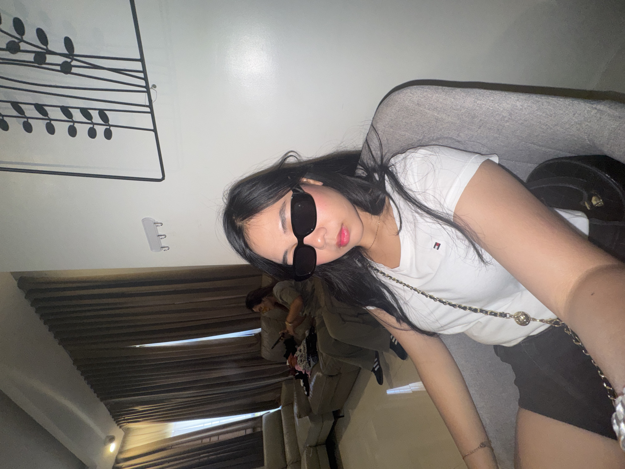 The image captures a woman wearing sunglasses and a white t-shirt, seated in a gray armchair. She is in the foreground, with her body angled towards the right side of the frame. Behind her, a brown sectional sofa is visible, occupied by another woman who is looking at a phone. Laundry is piled on the sofa. The environment appears to be an indoor living space, with brown curtains and a white wall adorned with a decorative metal art piece featuring vertical lines and circular shapes. The lighting is ambient, suggesting it is daytime. The location is identified as Davao City, Philippines.