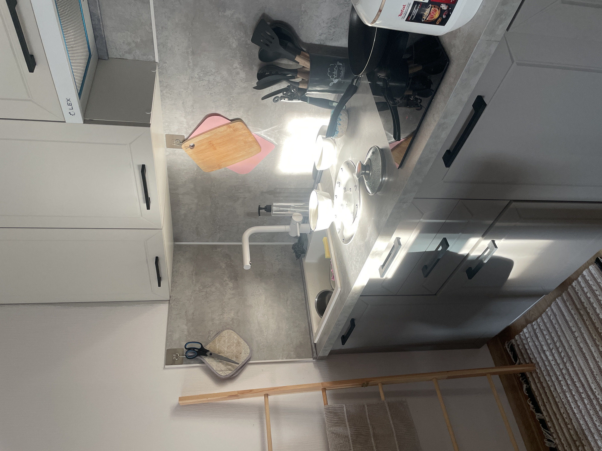 A sunlit, modern kitchen in Poroshkino, Russia, features a grey countertop with a sink containing a metal pot and a yellow sponge. White dishes and bowls are stacked on a plate. To the right, a black utensil holder overflows with cooking tools, including spatulas and tongs. A black frying pan rests on an induction cooktop next to a white Tefal appliance. Above the counter, white cabinets with black handles line the wall. A grey backsplash with a concrete texture extends behind the sink. On the left, a wooden towel rack holds beige towels, and a small striped rug lies on the floor. A pair of scissors hangs from a hook on the backsplash, next to two cutting boards. The scene is illuminated by bright sunlight streaming in, casting long shadows across the kitchen.