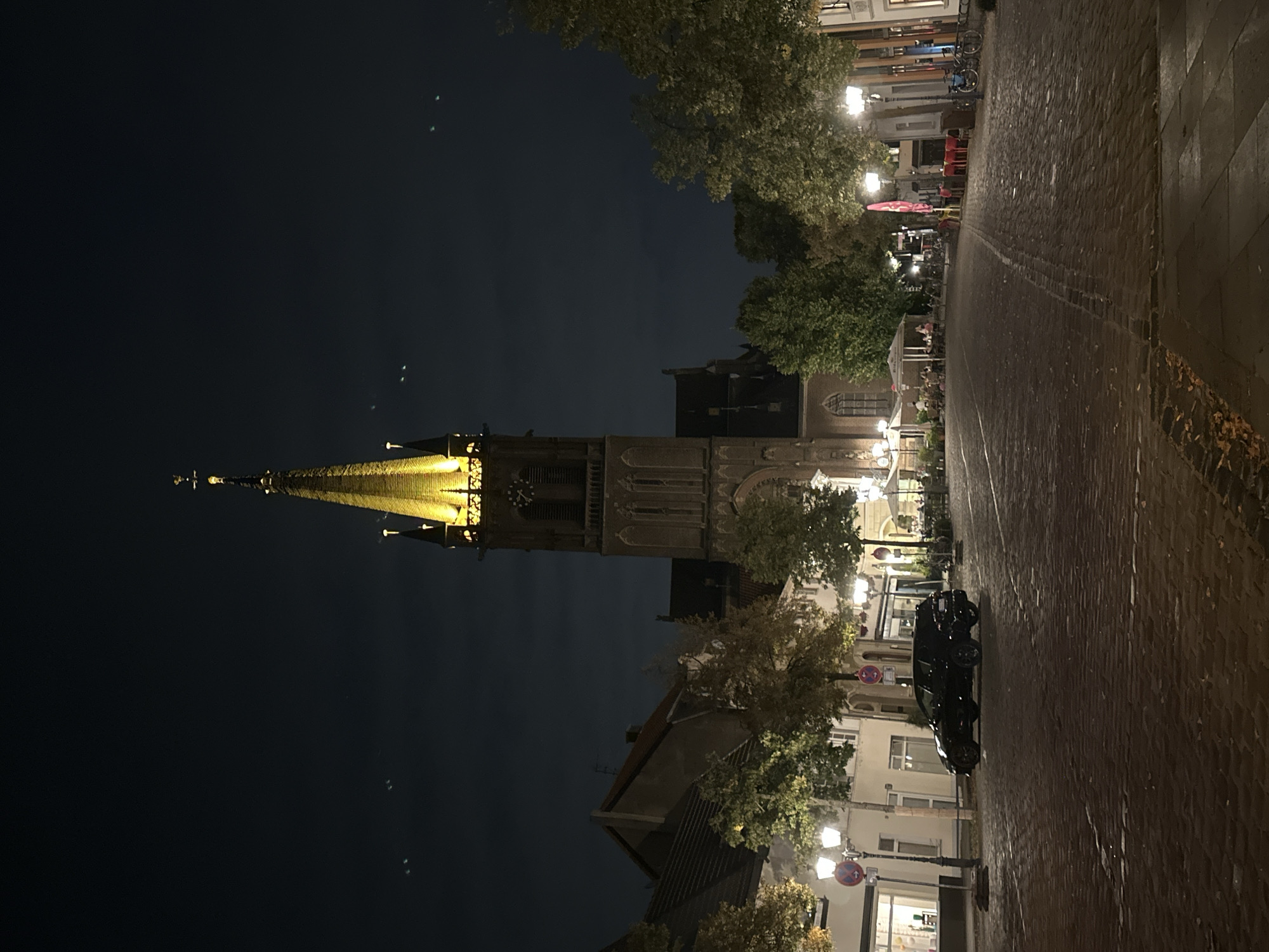 The image depicts a nighttime street scene in Krefeld, Germany, with a prominent church tower illuminated by artificial lights. The street is paved with cobblestones and lined with buildings and trees. A black SUV is parked on the left side of the street. In the background, there are several outdoor seating areas with umbrellas and chairs, suggesting a commercial or restaurant district. Streetlights are visible along the sidewalk. The sky is dark, with some faint clouds and what appear to be multiple small lights or objects scattered across it.