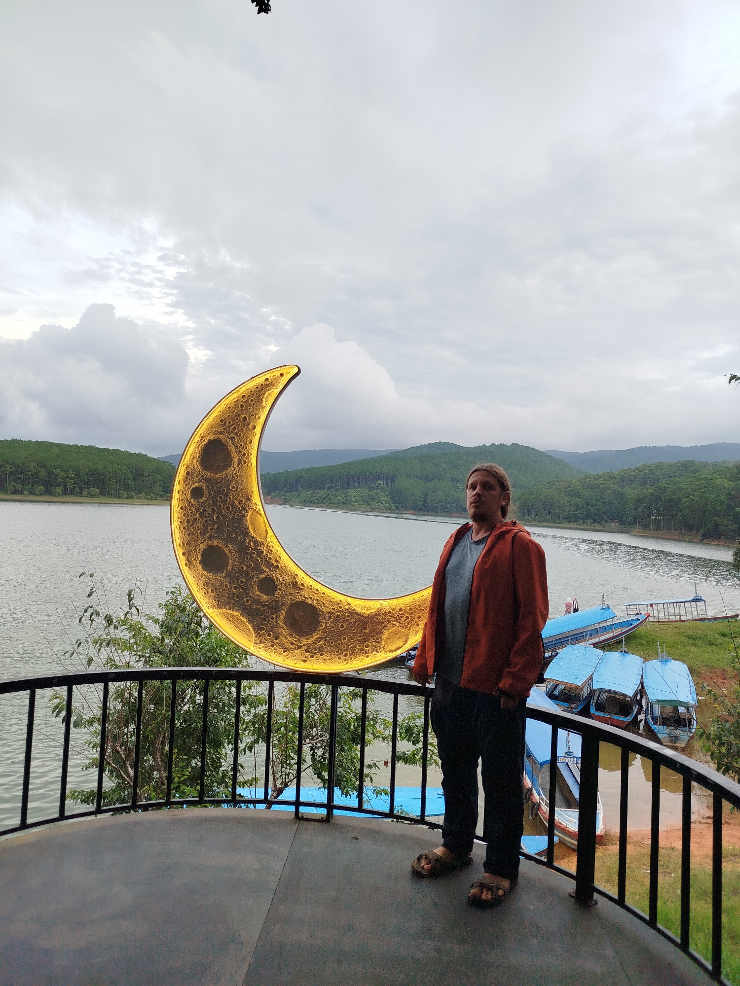 A man stands on a balcony overlooking a lake in Da Lat, Vietnam. He is wearing a rust-colored jacket over a grey t-shirt and dark pants. A large, illuminated crescent moon decoration is positioned to his left, partially obscuring the view of the lake. The lake is calm and reflects the overcast sky, which is filled with clouds. Dense green trees cover the hills that surround the lake, creating a lush backdrop. Several blue boats are docked along the shore. The balcony has a black metal railing. The time of day appears to be late afternoon or early evening, given the subdued lighting and cloudy weather. The man has a neutral expression.