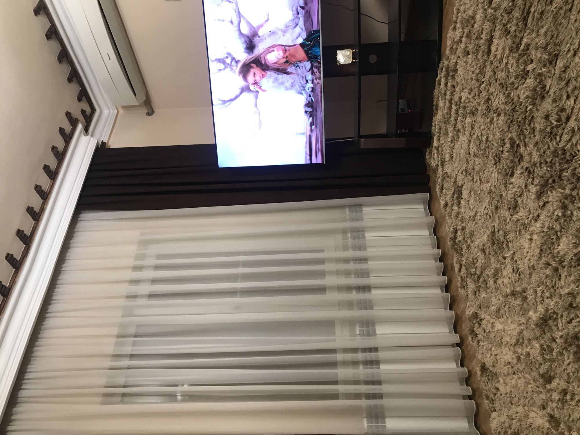 A television is mounted above a window in a room in Yerevan, Armenia. The television screen displays a fair-skinned woman with long brown hair, wearing a green and black outfit, partially obscured by a digital tree or foliage, with a bright light source in the background. A dark brown valance covers the top of the window, which has layered white shades with horizontal slats. To the left, a white ceiling with ornate brown and white trim is visible. To the right, part of a black television stand and a beige shag rug are visible.