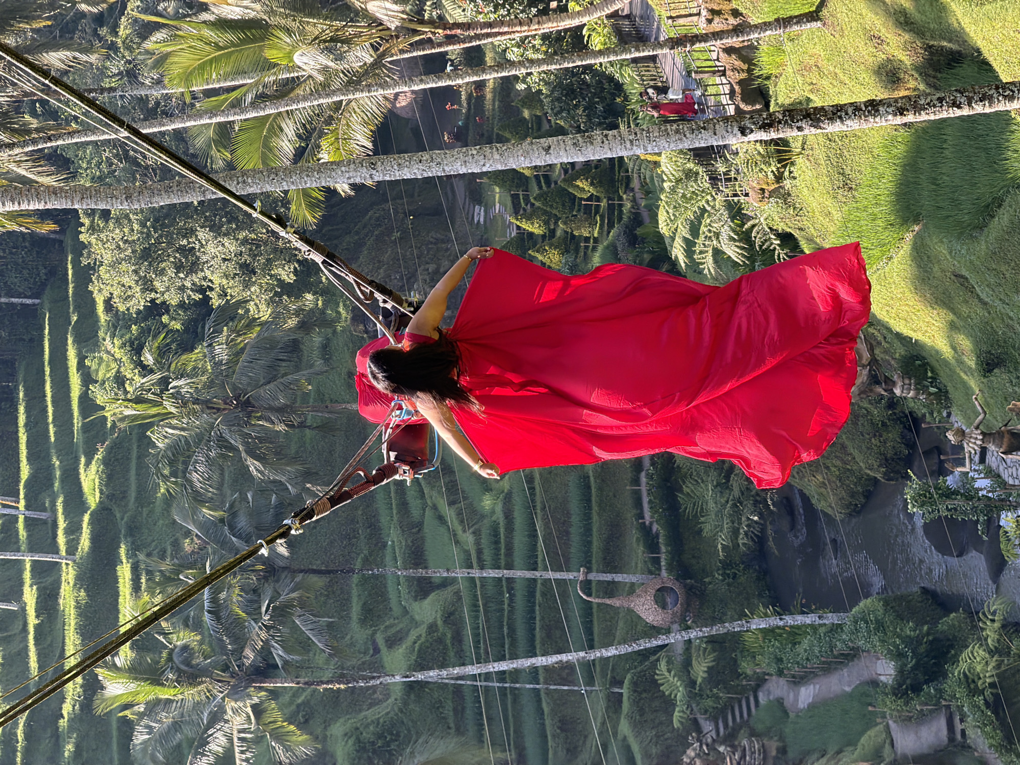 In Tegallalang, Indonesia, a woman in a vibrant red dress is captured mid-swing on a high jungle swing. She is viewed from behind, her arms outstretched, the voluminous fabric of her dress billowing around her. Below and beyond, the iconic Tegallalang rice terraces cascade down the valley, a lush green tapestry interspersed with palm trees. A narrow river snakes through the scene, and pathways are visible winding through the terraces. In the background, other tourists can be seen on a wooden walkway, some taking photographs. The lighting suggests it is daytime, with sunlight illuminating the vibrant greens of the landscape and casting shadows.