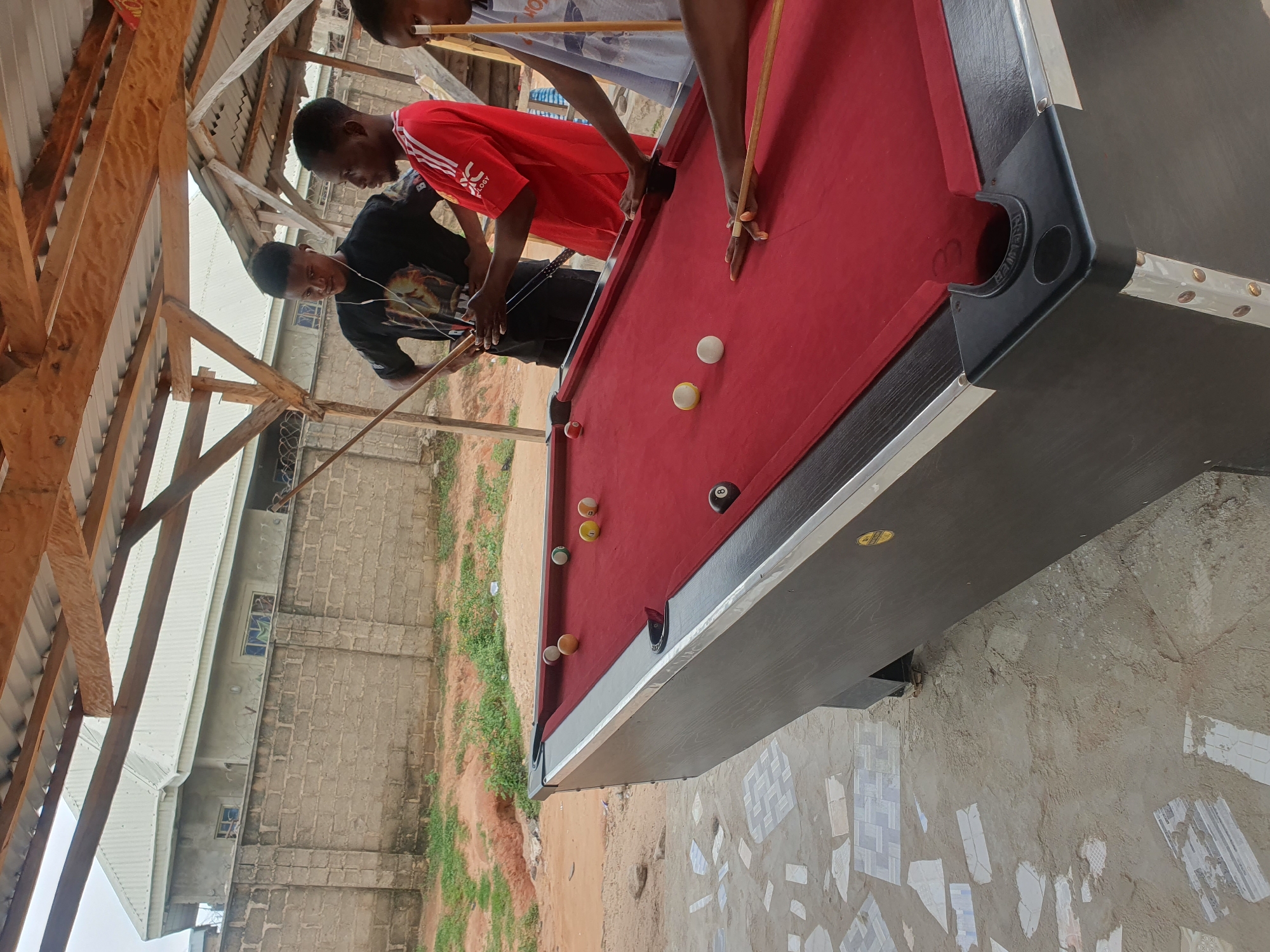 In Lokoja, Nigeria, four young men are gathered around a red-felted billiard table under a makeshift wooden and corrugated metal roof. The scene appears to be an outdoor recreational area, possibly a local hangout or bar, with a brick wall and an unfinished building visible in the background. The ground is a mix of dirt, sand, and scattered broken tiles, suggesting an undeveloped or in-progress construction site.
One man, wearing a black t-shirt with a graphic print and white earphones, holds a billiard cue and seems to be waiting for his turn. Another man, in a red Adidas t-shirt, is actively engaged in the game, leaning over the table with a cue in hand. A third man, in a light-colored t-shirt, stands behind the red-shirted man, also holding a cue. The fourth individual is partially visible to the right, holding a cue and facing the table.
Several billiard balls are scattered on the red felt: a solid yellow ball, a white cue ball, a striped yellow ball, a white solid ball, a green-striped ball, and the black eight ball. The billiard table itself has a black frame with silver trim and black corner pockets. The overall atmosphere suggests a casual and relaxed gathering focused on playing billiards.