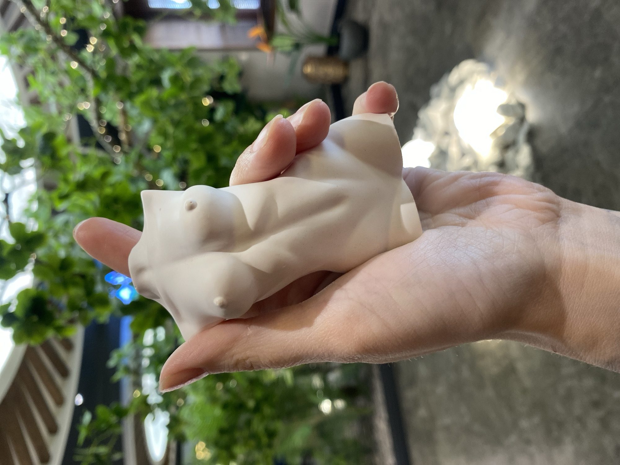 A close-up shot features a person's hand holding a small, white, abstract sculpture that appears to depict a torso. The sculpture is cradled in the palm, with the person's fingers gently curved around it. The background is blurred, but it suggests an indoor setting near a window or opening with natural light and foliage. There are green plants visible through the window. The location cannot be determined to be the city of Votkinsk, Russia, due to lack of any identifiable indicators.