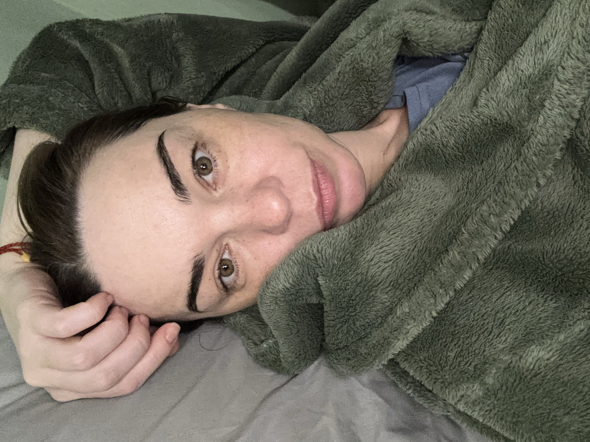 A woman is lying on a grey bed covered with a soft, green blanket. She is partially enveloped by the blanket, which is draped around her face and body. Her head is resting on her hand, and she is looking directly at the camera. Her features are visible, including her eyebrows, eyes, nose, and lips. Her hair is dark. The background is blurred and indistinct. The location is Unknown, Unknown.