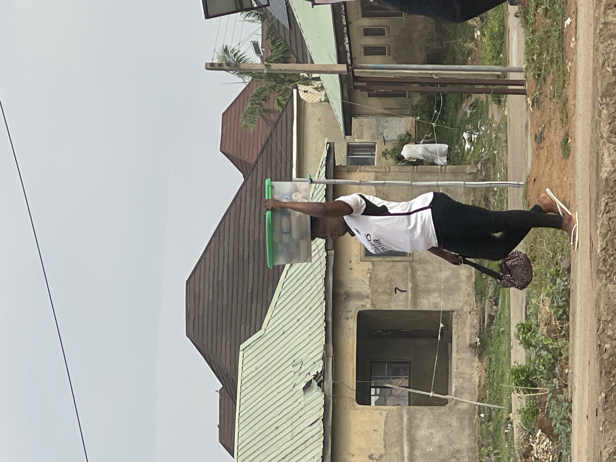 An individual is depicted standing outdoors on an unpaved road or path in what appears to be a residential area of Abuja, Nigeria. The person is balancing a clear plastic container with a green lid on their head, a common method of carrying goods in the region. They are dressed in a white short-sleeved shirt with black accents on the sleeves and black pants, complemented by white flip-flops. A dark, patterned bag is held in their left hand, hanging low.
The background features multiple residential buildings under an overcast sky. Some structures have corrugated metal roofs, with one appearing brown and another light green or grey. Other buildings are made of concrete, showing visible windows and open doorways. Several utility poles, one equipped with a small solar panel, are present. Sparse green vegetation, including bushes and grass, grows amidst the buildings and along the unpaved ground.
The image suggests daytime, characterized by an overcast or cloudy sky with no strong shadows. On the individual's shirt, partially visible text reads "BREAD OF LIFE MINISTRIES". A faint number "7" can be seen on the wall of one of the concrete buildings in the background.