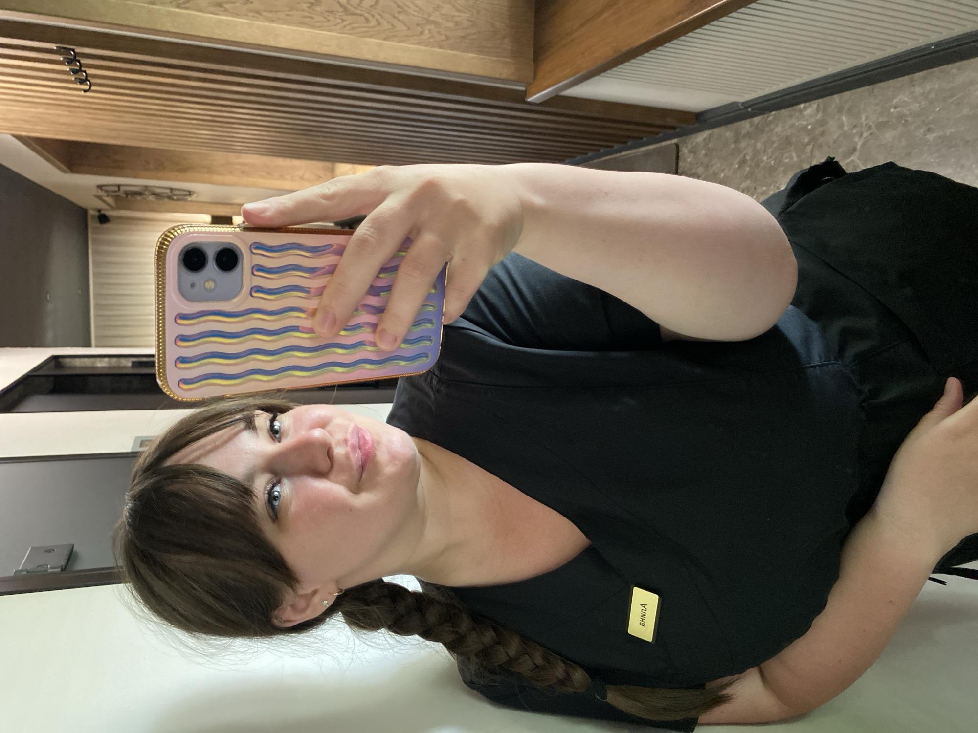 A woman takes a selfie in an indoor location that appears to be Votkinsk, Russia. She is wearing a black uniform with a name tag and has a braid. She is holding a phone with a pink case that has a wave pattern. The background includes light colored walls, a ceiling with wooden beams, and a marble countertop.