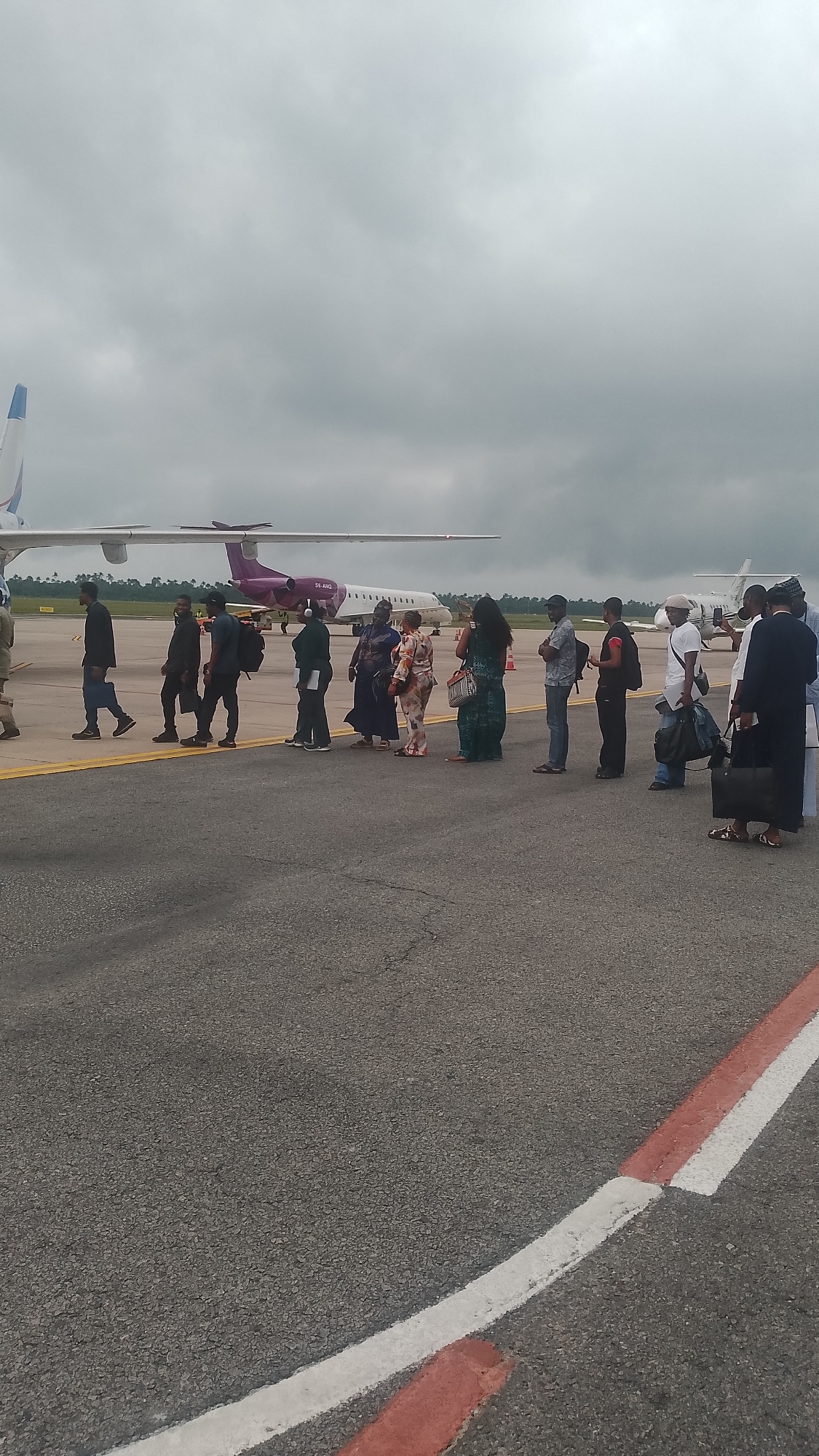 A group of approximately 15 people are lined up on an asphalt tarmac, standing in front of a parked airplane. The airplane, a narrow-body jet with purple accents and the registration "5H-AXQ," is positioned behind the individuals. The sky is overcast with gray clouds. The people are dressed in various casual and semi-formal attire. They appear to be boarding or disembarking the aircraft. The scene suggests an airport environment, possibly at a regional airport in or near Orerokpe, Nigeria, given the presence of a commercial airplane.