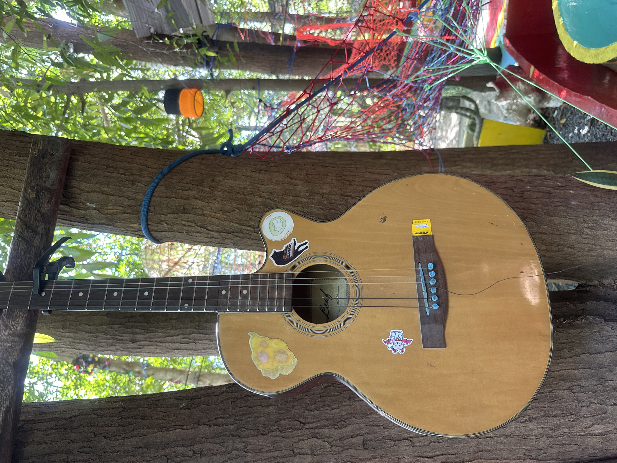 A light-colored acoustic guitar with several stickers affixed to its body rests against a tree trunk. The guitar has a capo on its neck and appears to be strung. Above the guitar, multicolored decorations hang from the tree branches. The location is likely in ตำบลเกาะพะงัน, Thailand.