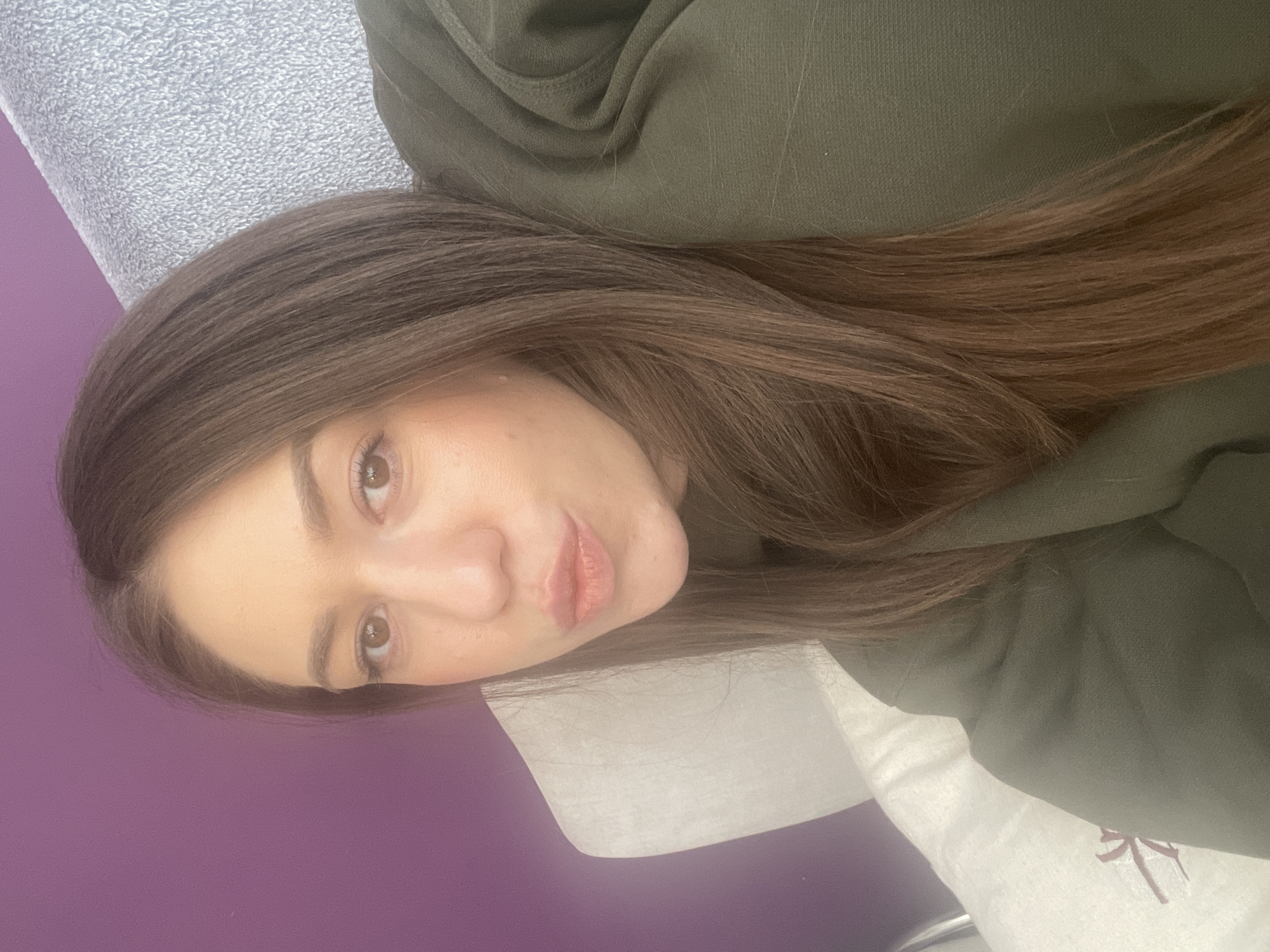 A selfie shows a woman with brown hair wearing an olive green hoodie. She is lying on a surface covered with a white cloth or pillow, and behind her is a purple-colored wall. The location is impossible to ascertain based solely on visual features of the image.
