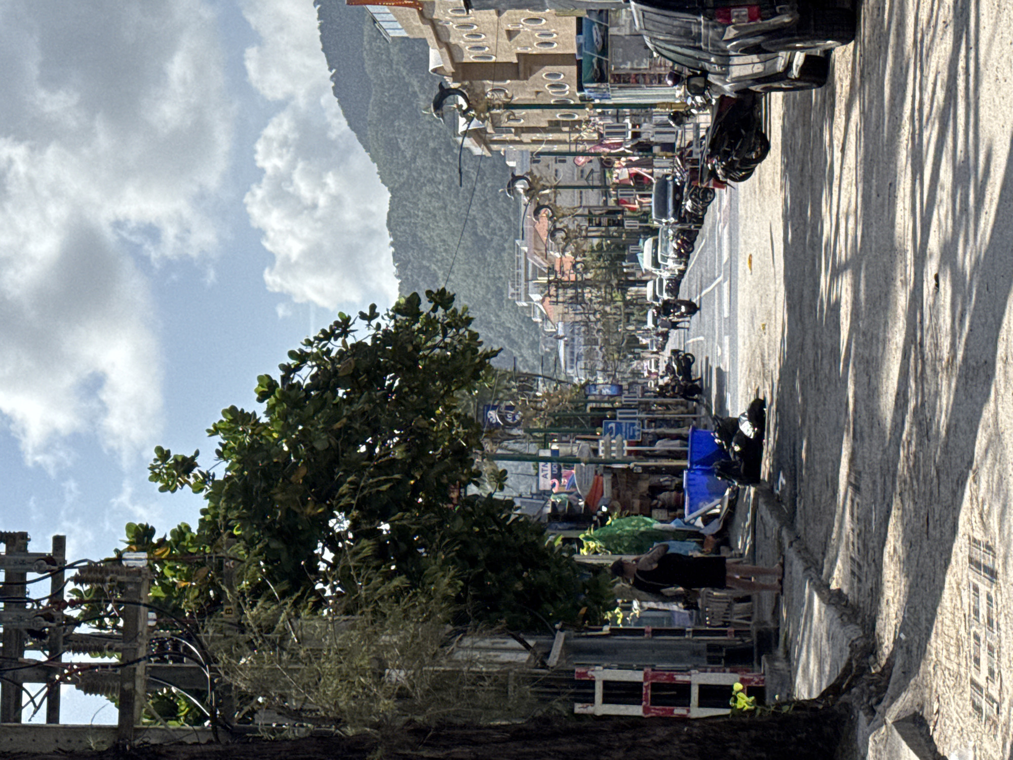 A street scene in Patong, Thailand, shows a moderately busy street lined with buildings and a mountain in the background. On the left side of the street, a man in a black tank top and shorts stands on the sidewalk, facing away from the camera. A large tree with green leaves is also on the left, casting shadows on the ground. The street is paved with asphalt and has various vehicles, including cars, motorcycles, and scooters, parked on the sides and driving down the road. Some of the buildings appear to be shops or restaurants, with signs and awnings visible. The sky is partly cloudy, with a mix of sunlight and shadows creating a dynamic lighting effect on the scene. The overall atmosphere is one of a typical day in a tourist area, with a blend of local life and visitor activity.