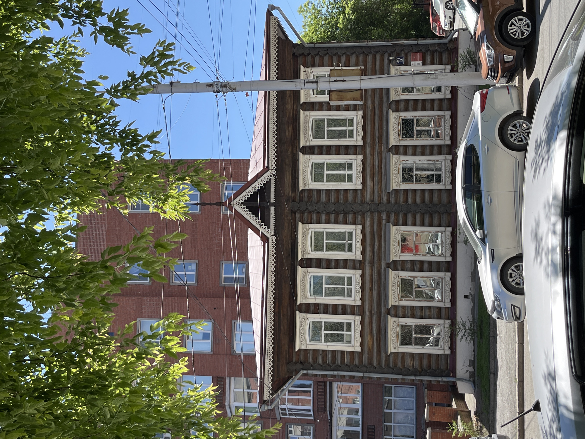 The image depicts a scene dominated by a traditional wooden house with ornate window frames, set against a backdrop of a modern brick building and trees. The wooden house, constructed from logs, features several windows with decorative white trim, suggesting a historical architectural style. Adjacent to the wooden house is a multi-story brick building with numerous rectangular windows. Green foliage from trees partially obscures the view of both buildings, particularly in the foreground and upper left. A clear blue sky is visible above.
Several cars are parked along the street in front of the buildings, indicating an urban or semi-urban setting. These include a white sedan, a brown SUV, and a portion of another car. Power lines crisscross the sky, a common sight in many developed areas. One of the wooden house's windows has a small sign with the number "2" on it. The lighting suggests it is daytime, likely a sunny day, given the bright illumination and shadows. The overall scene appears peaceful and ordinary, showcasing a juxtaposition of old and new architecture. No people are visible in the image.