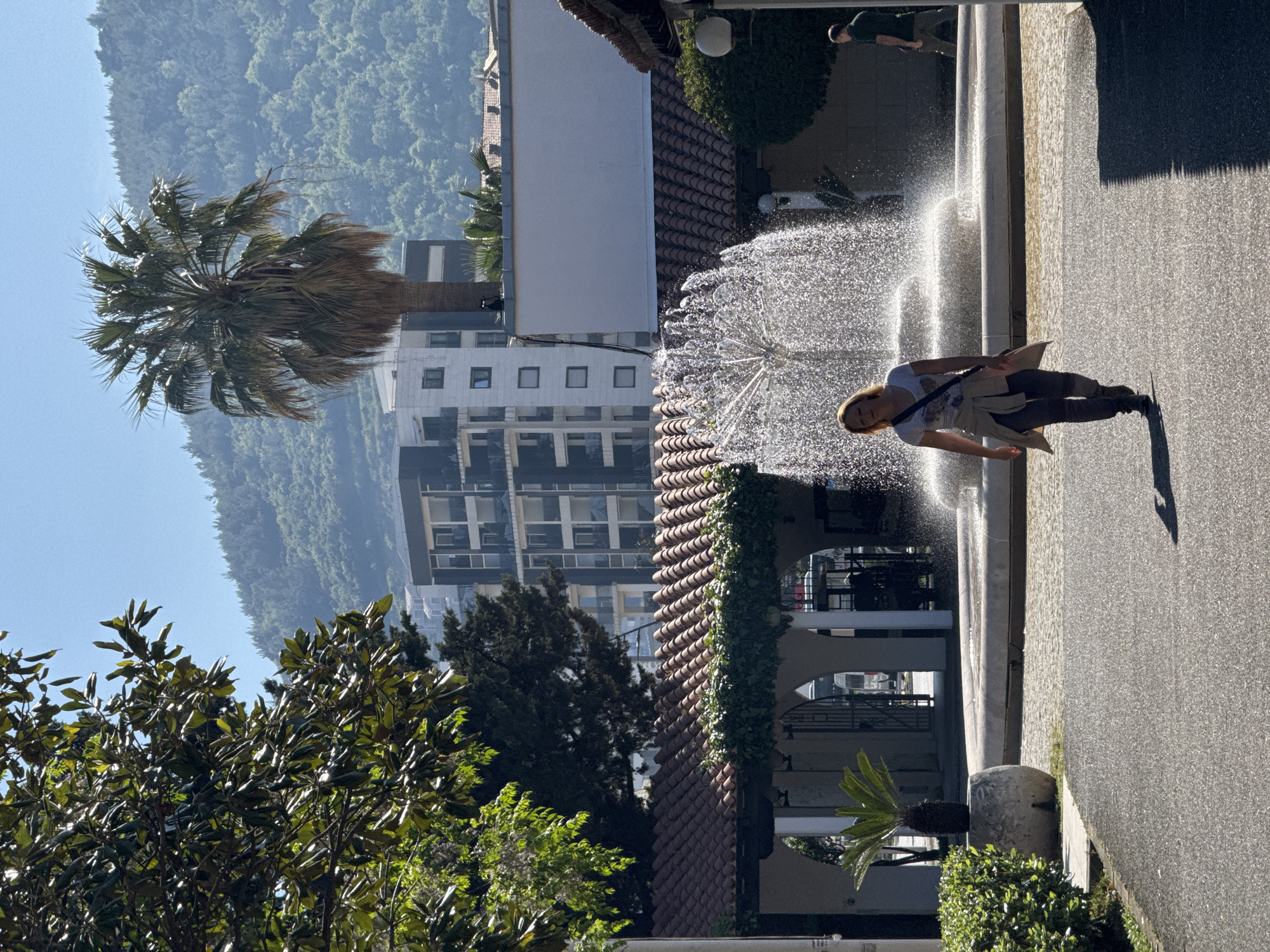 This image, captured in Budva, Montenegro, presents a sunlit outdoor scene featuring a prominent water fountain, a person, and a mix of natural and architectural elements. In the mid-ground, a woman with light-colored hair stands on a paved surface, facing towards an active fountain. She is dressed in a light t-shirt, dark trousers, and has a light jacket tied around her waist, carrying a dark shoulder bag. The fountain vigorously sprays water in various directions, creating a sparkling effect under the bright sunlight. A second person, dressed in dark clothing, is partially visible further back beyond the fountain's spray.
To the left of the fountain, a large palm tree stands tall, its fronds catching the sunlight. Lush green foliage, including various trees and bushes, is present in the foreground, mid-ground, and background. The architectural landscape comprises buildings with distinctive reddish-brown tiled roofs alongside more modern, multi-story structures featuring numerous windows.
In the distant background, a large, densely forested mountain or hillside rises against a clear, bright blue sky, underscoring the natural beauty of the region. The strong sunlight suggests it is midday or early afternoon, casting distinct shadows across the scene. The overall atmosphere is bright and pleasant, characteristic of a sunny day in a coastal resort area. No visible text is present in the image.