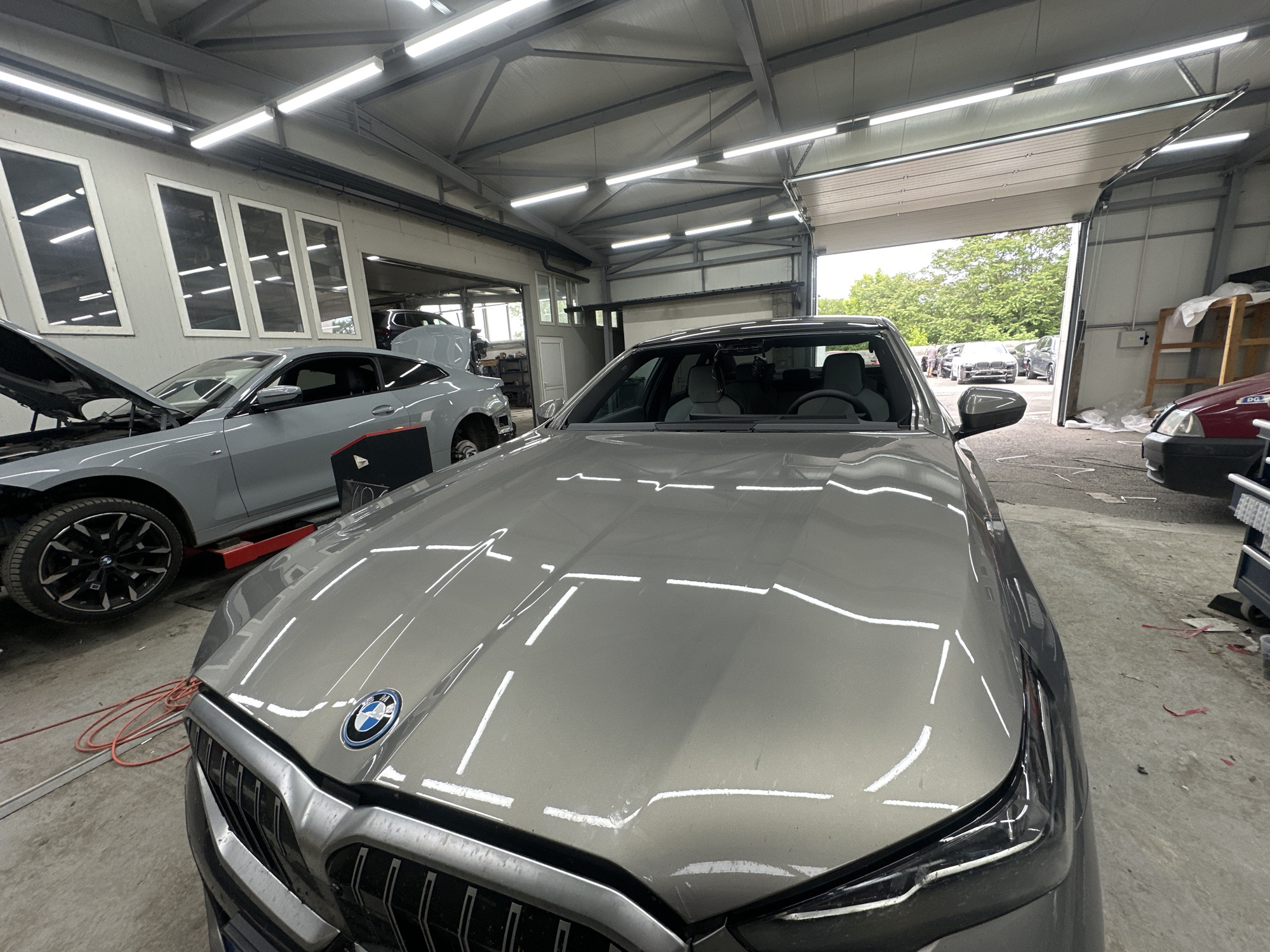 The image shows the front of a gray BMW parked inside what appears to be an auto repair shop. The car is prominently positioned in the foreground, with its hood taking up much of the view. A portion of another gray car with its hood open is visible to the left. The repair shop has multiple garage doors, one of which is open, revealing an outdoor scene with other vehicles. The shop is lit by fluorescent lights. An orange extension cord is lying on the floor near the BMW.