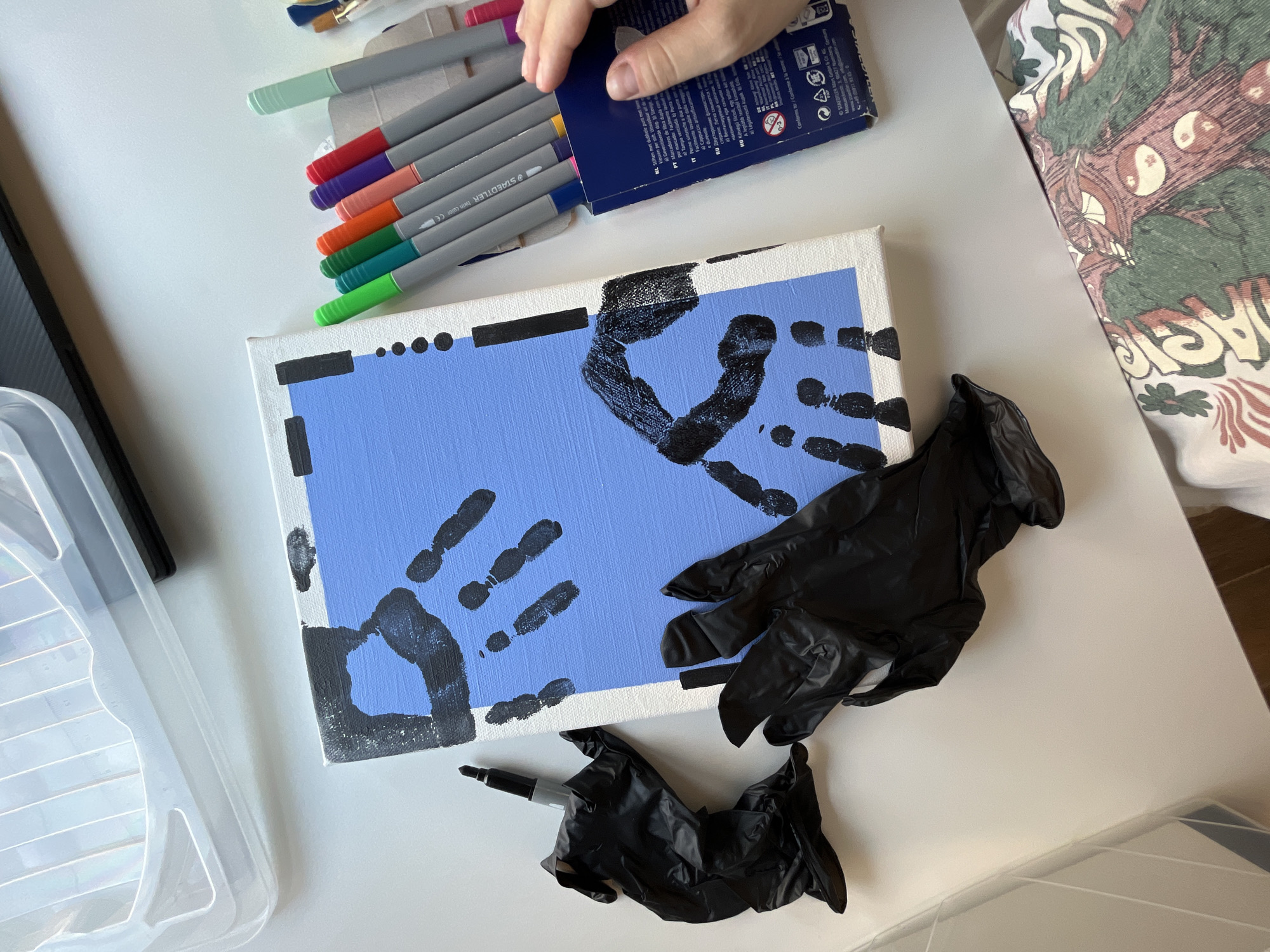 A top-down, eye-level, medium shot captures a white table surface with art supplies. The primary subject is a small, white canvas, approximately 8x10 inches, partially painted with a light blue background. Black paint is applied in the shape of handprints on the canvas. To the left of the canvas are two crumpled black disposable gloves and a black marker. To the right of the canvas is a blue cardboard box holding an array of colorful Staedtler brand markers, several of which are visible in the box and a human hand is reaching into the box. A clear plastic container with compartments is visible in the upper left corner of the frame. The background consists of a white tabletop with a sliver of what appears to be a patterned fabric in the bottom right corner. The scene depicts an art creation process. The image does not provide any specific information about the location in the city of Limassol, Cyprus.
