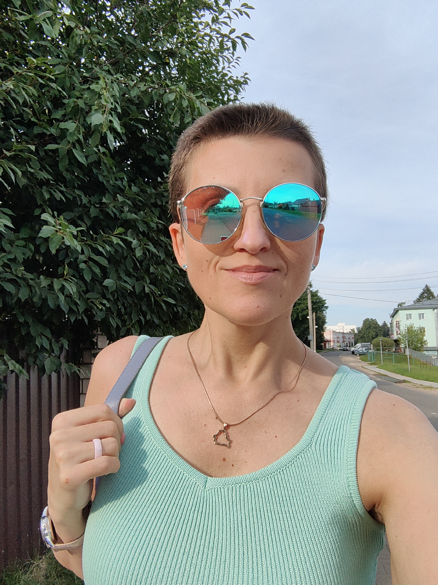 A woman with short brown hair and sunglasses stands outdoors, facing the camera. She is wearing a mint green tank top and a gold necklace with a pendant shaped like the territory of Belarus. A gray strap from a bag is visible over her left shoulder. On her left hand, she wears a light pink ring. A brown wooden fence is behind her to the left, and a tree with green leaves is to her left and behind her. To her right, a street with buildings and utility poles is visible. The sky is overcast.