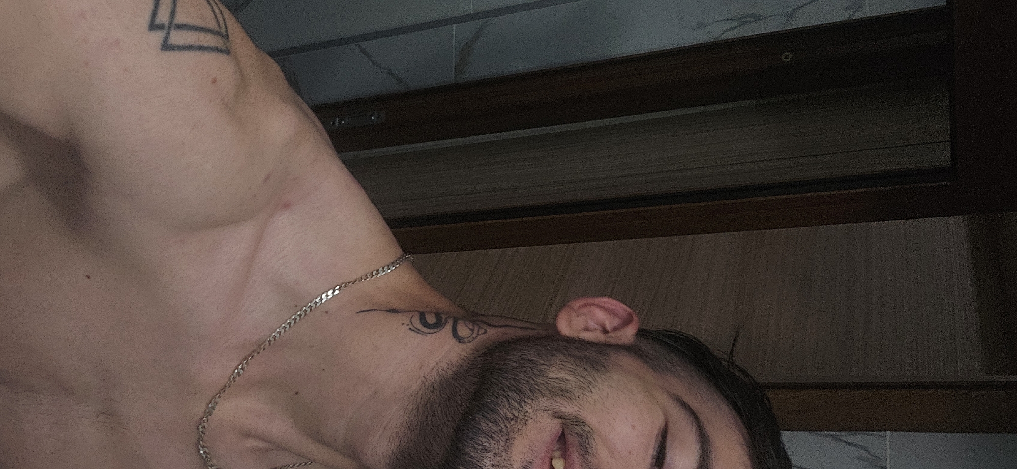 A close-up, low-angle shot depicts a man's upper body and face. He has a short beard and dark hair. Visible on his skin are tattoos; one on his chest/shoulder area and another near his neck. He is wearing a thin silver chain necklace. The background is composed of a dark brown wooden structure with light-colored paneling above and white marble-like tiling on top. The location within Plovdiv, Bulgaria, is not discernible from the image.