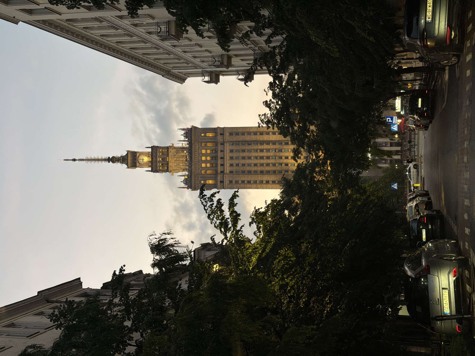 The image shows the Palace of Culture and Science in Warsaw, Poland, viewed from a street perspective. The sky is pale with light clouds, suggesting late afternoon or early evening. Buildings flank the palace on either side, with the streetscape partly obscured by trees. Parked cars line the right side of the street, and some pedestrians are visible further down the road near crosswalk signs. The architecture of the palace is illuminated, highlighting its tower and clock faces.