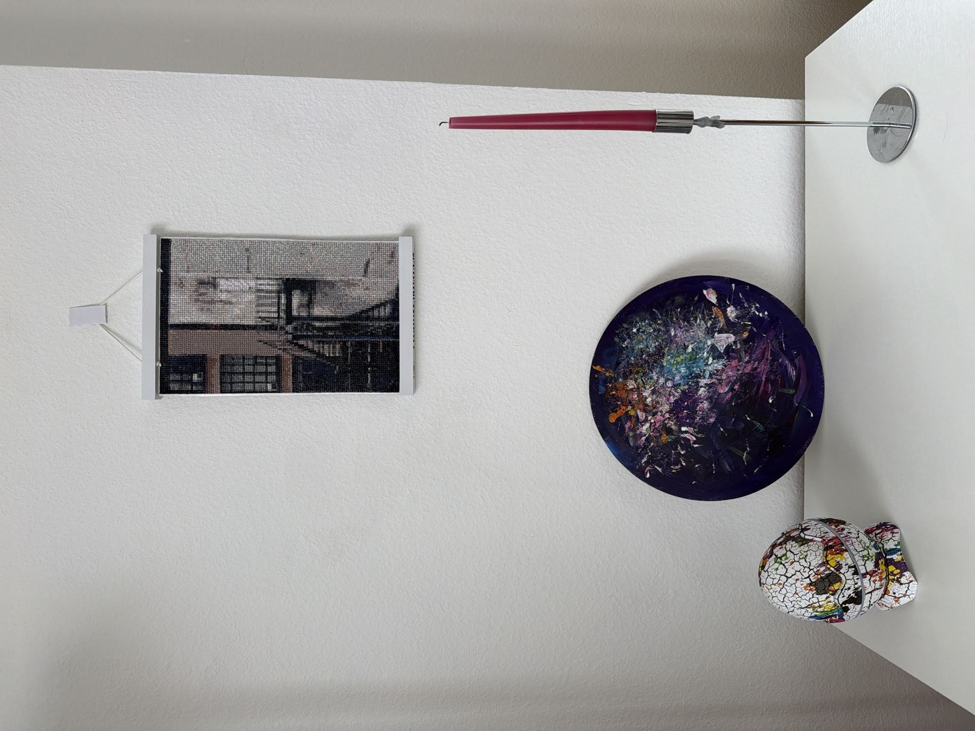 A low-angle, brightly-lit indoor shot shows a white wall decorated with a framed picture, a red reading lamp, an abstract circular painting, and a decorative fish sculpture.
The framed picture on the left, mounted with adhesive strips, features a grayscale cityscape. Above it, a thin red reading lamp extends from the wall towards the right with a silver fixture.
To the right and slightly below the lamp is a round painting dominated by dark blues and purples with splashes of white, orange, and pink.
Beneath the painting, placed on the edge of a white cabinet or column, is a decorative fish sculpture. The sculpture is white with a cracked texture and filled with colorful patterns.
The wall has a slightly textured surface. The setting appears to be a modern interior.