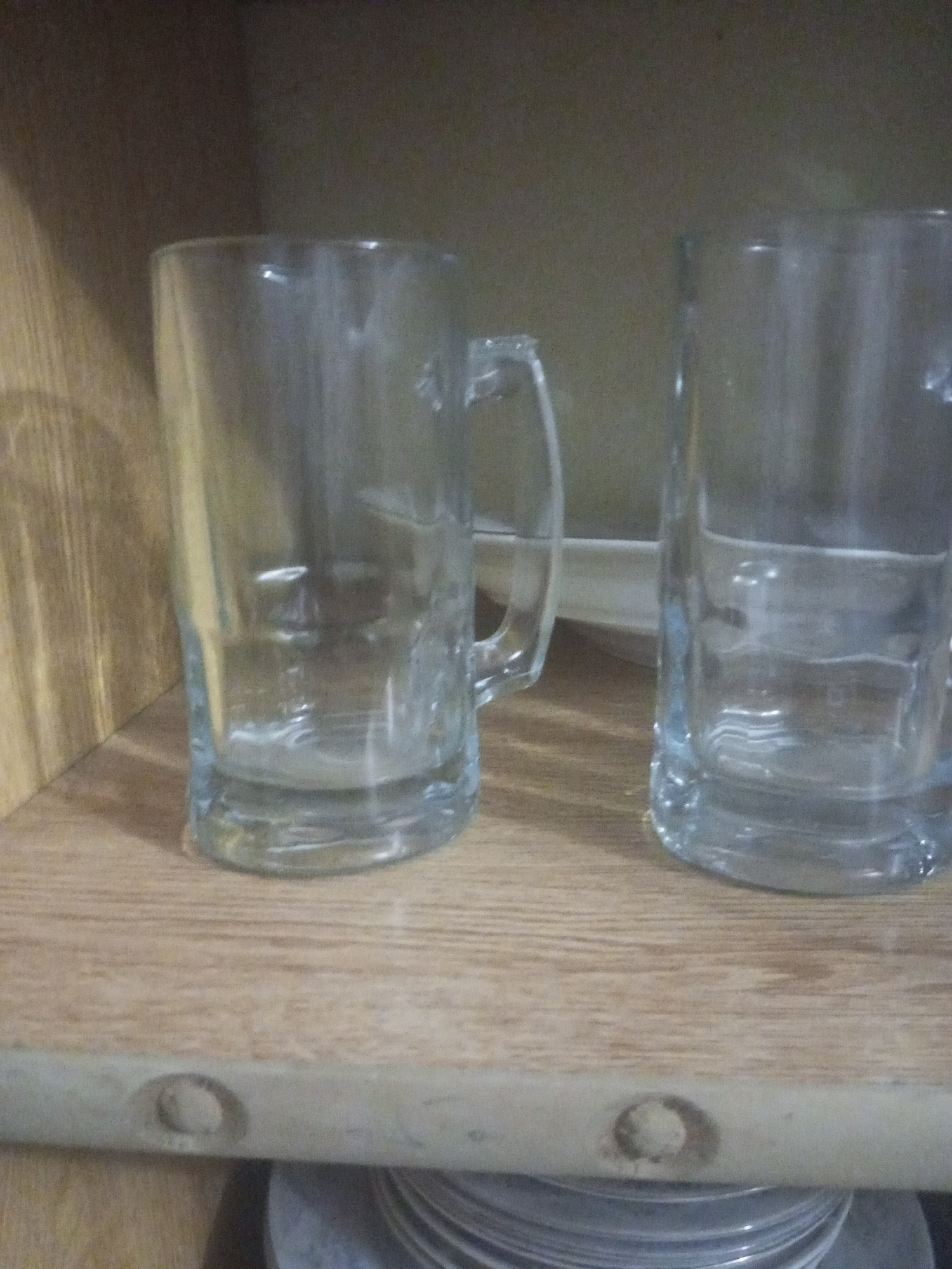 Two empty glass mugs sit on a wooden shelf. The mug on the left has a handle. The shelf is part of a cabinet. The background is dark and out of focus. The scene appears to be indoors. The specific location in Mansehra, Pakistan is not discernible from the image.