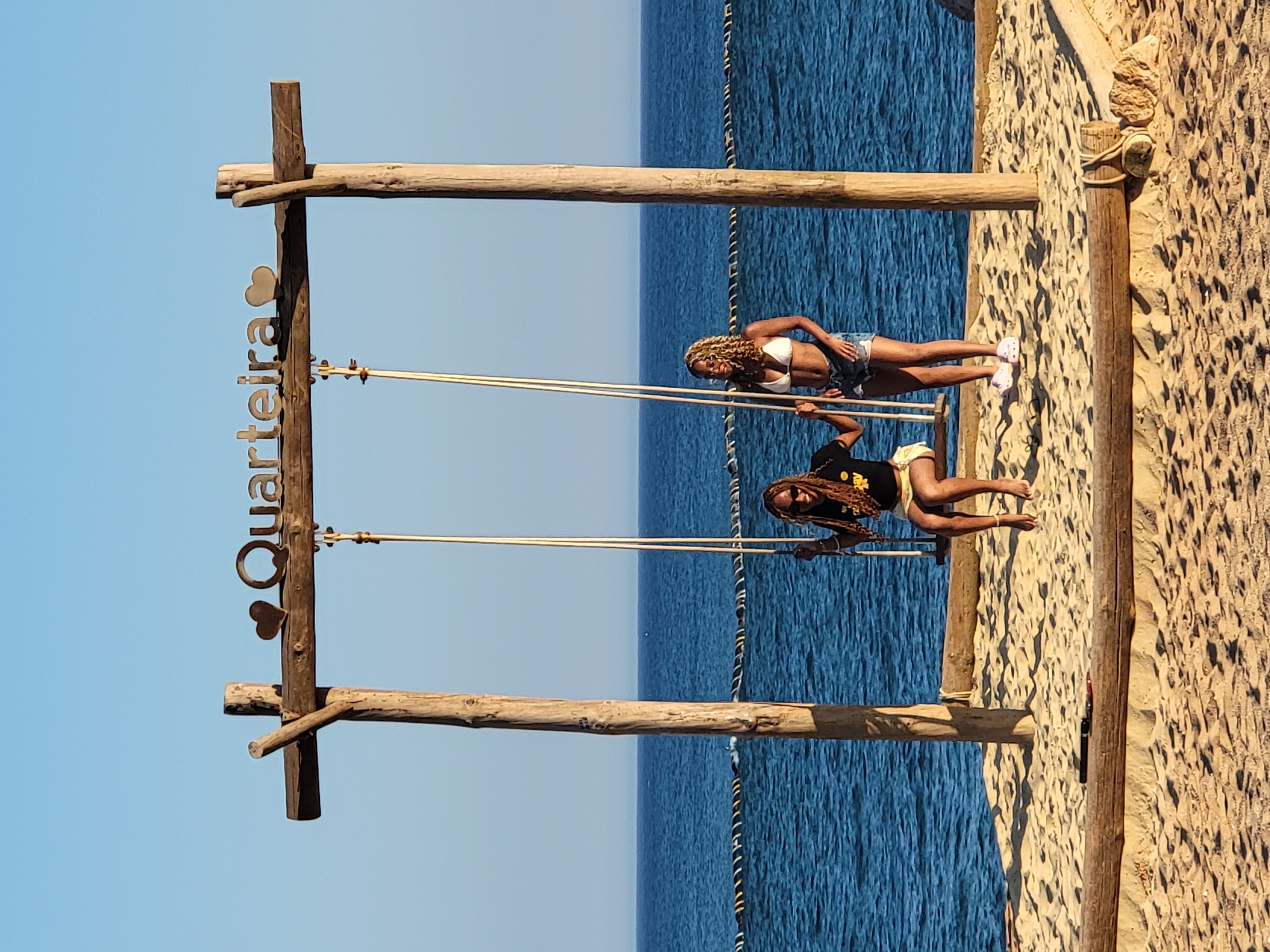 Two women are standing near a swing set on a sandy beach. The woman on the left is sitting on the swing and wearing a black t-shirt with yellow writing and yellow shorts. She has long, braided hair and is wearing sunglasses. The woman on the right is standing next to the swing and wearing a white bikini top and denim shorts. She has curly blonde hair and is wearing glasses. A wooden sign above the swing set reads "Quarteira" with two heart shapes. The ocean is visible in the background with a clear blue sky. The beach is made of sand with some wooden logs and ropes bordering it.