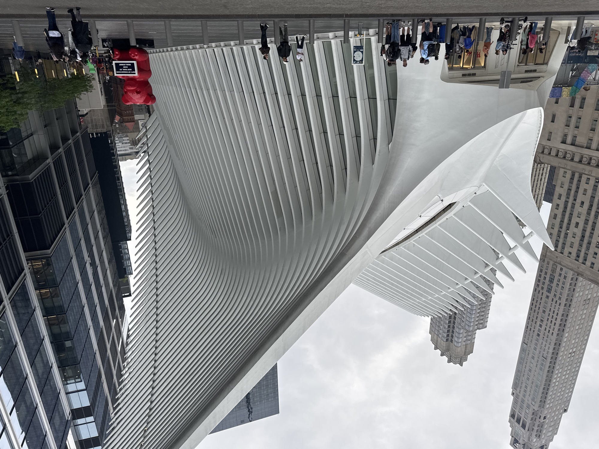 The image shows the Oculus, a transportation hub and shopping mall located in the World Trade Center complex in Lower Manhattan, New York City. The Oculus has a distinctive white, winged exterior design. People are walking along the perimeter of the building. Adjacent to the Oculus are tall modern skyscrapers. A large, red bear sculpture stands along the pedestrian area. The sky is overcast.