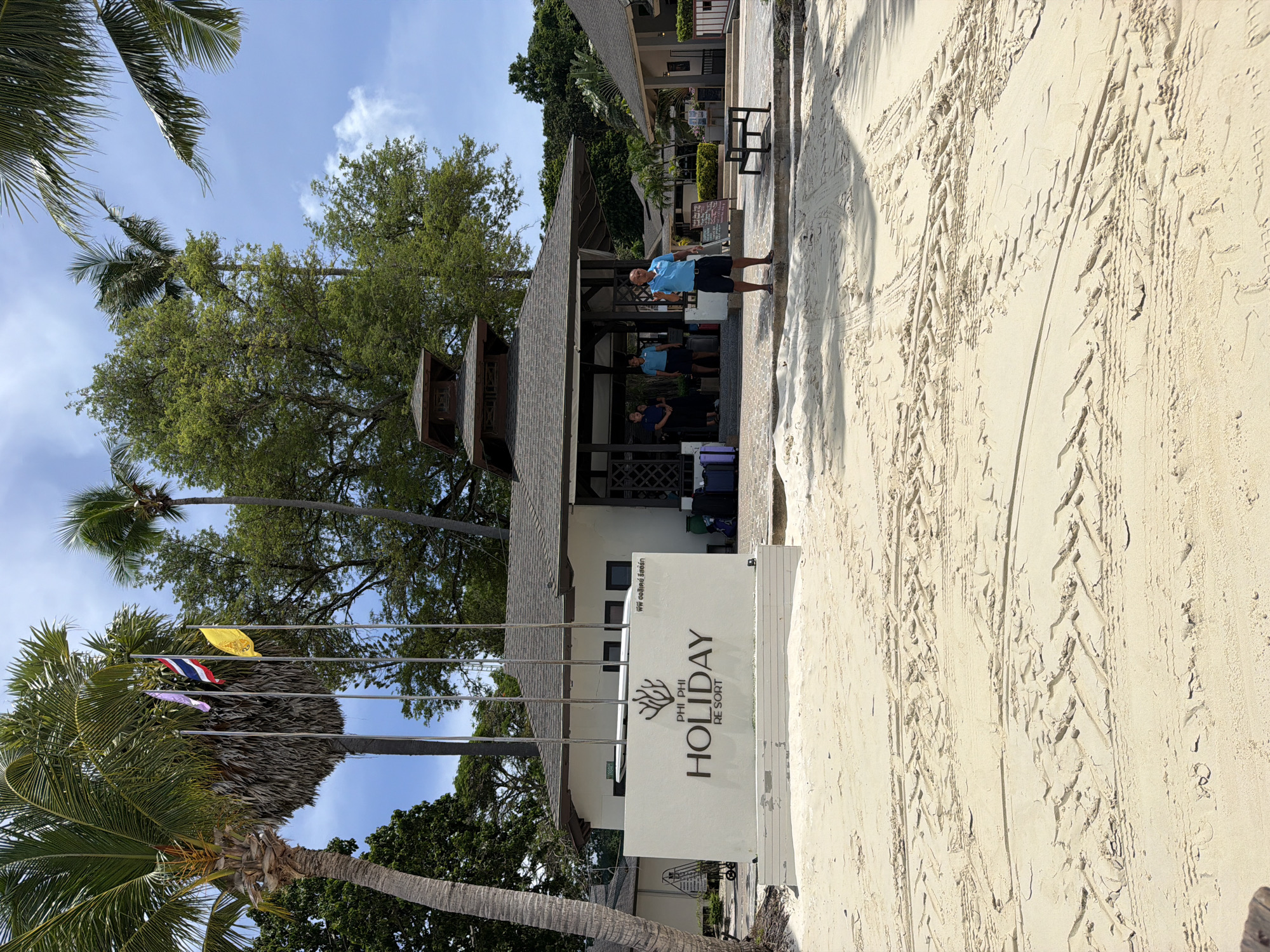 The image depicts the entrance to the Phi Phi Holiday Resort in Ao Nang, Thailand. The foreground is a sandy beach with tire tracks imprinted on it. Behind the beach, a white sign reads "PHI PHI HOLIDAY RESORT" in large, dark lettering. The resort building features a light-colored facade with dark trim and a dark, multi-tiered roof. Several palm trees are visible, including a large one on the left and others to the right and in the background. A man in a blue shirt and dark shorts stands in front of the resort, gesturing with his right hand. Two women in blue shirts and dark skirts stand near the entrance of the building. Luggage, including suitcases, is placed near the entrance. Flags fly from poles near the palm tree on the left. The sky is blue with scattered clouds.