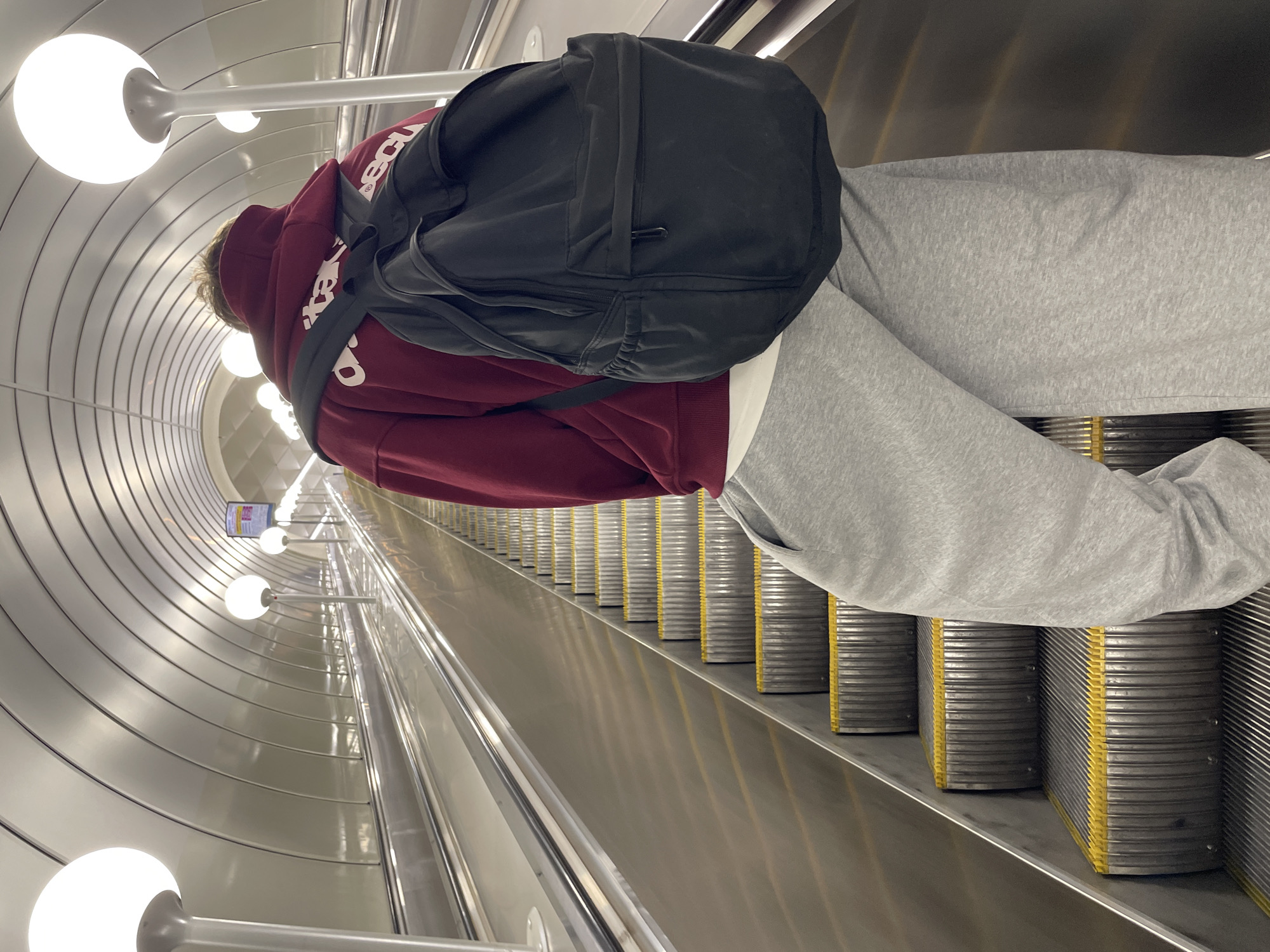 A person wearing a maroon hoodie, grey sweatpants, and a black backpack stands on an escalator in a metro station. The station's walls are curved and lined with evenly spaced, spherical lights mounted on poles. The escalator's steps are grey with yellow trim. The location is Saint Petersburg, Russia.