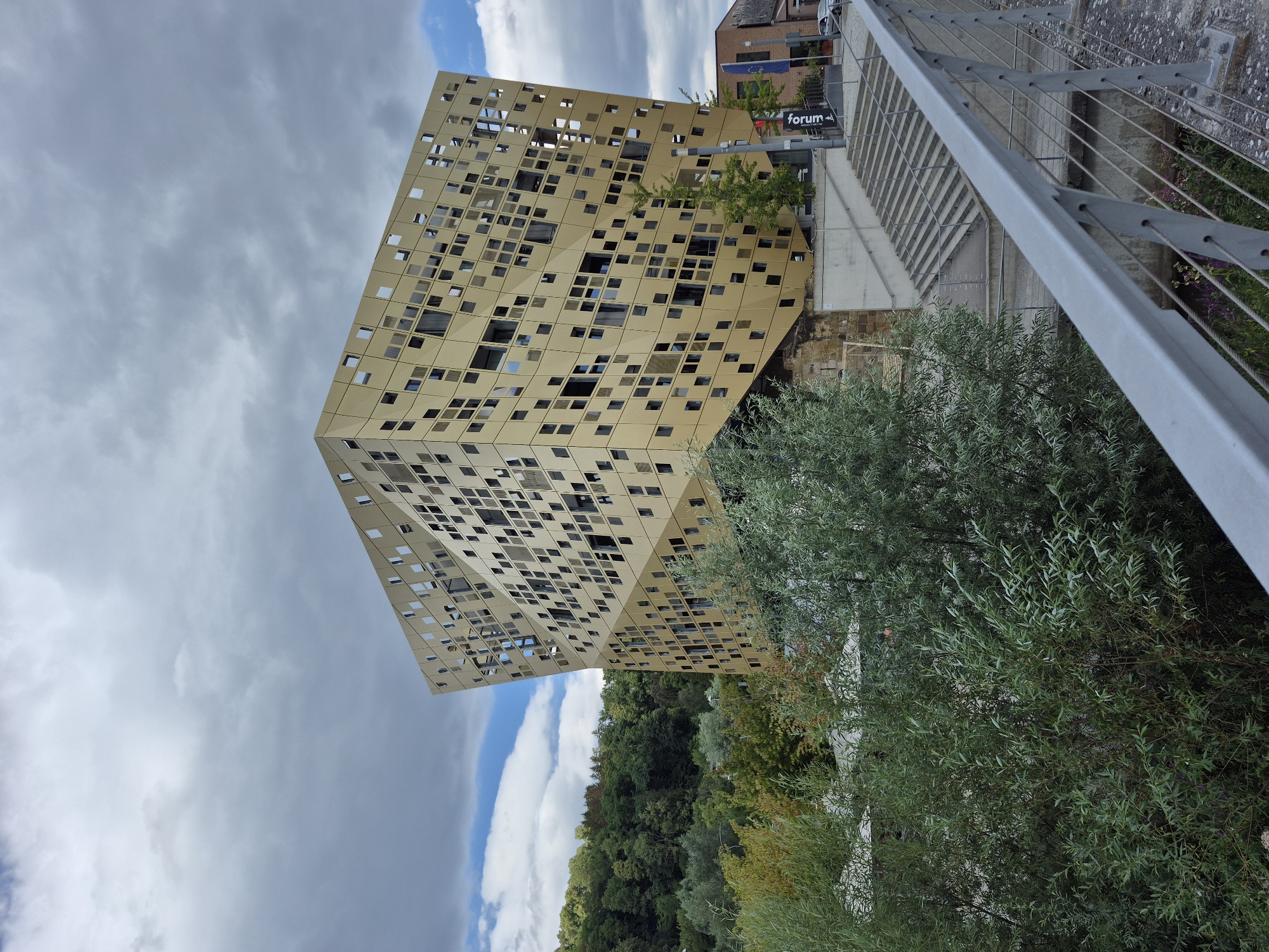 The image shows the Forum Gold und Silber, a striking modern building located in Schwäbisch Gmünd, Germany. The building's facade is a distinctive gold-colored material with a repeating pattern of square openings, giving it a unique, almost crystalline appearance. It is situated on a gentle slope, with a set of stairs leading up to an entrance area. Lush green trees and foliage are visible in the foreground and on the hillside behind the building, suggesting a park-like or natural setting within the city. The sky is partly cloudy, with a mix of grey clouds and patches of blue. A metal railing from what appears to be a bridge or elevated walkway is in the immediate foreground, angled into the scene. A flag and signage are visible near the building's entrance, identifying it as "Forum Gold und Silber". The overall impression is one of modern architecture integrated into a green, possibly riverside, environment.