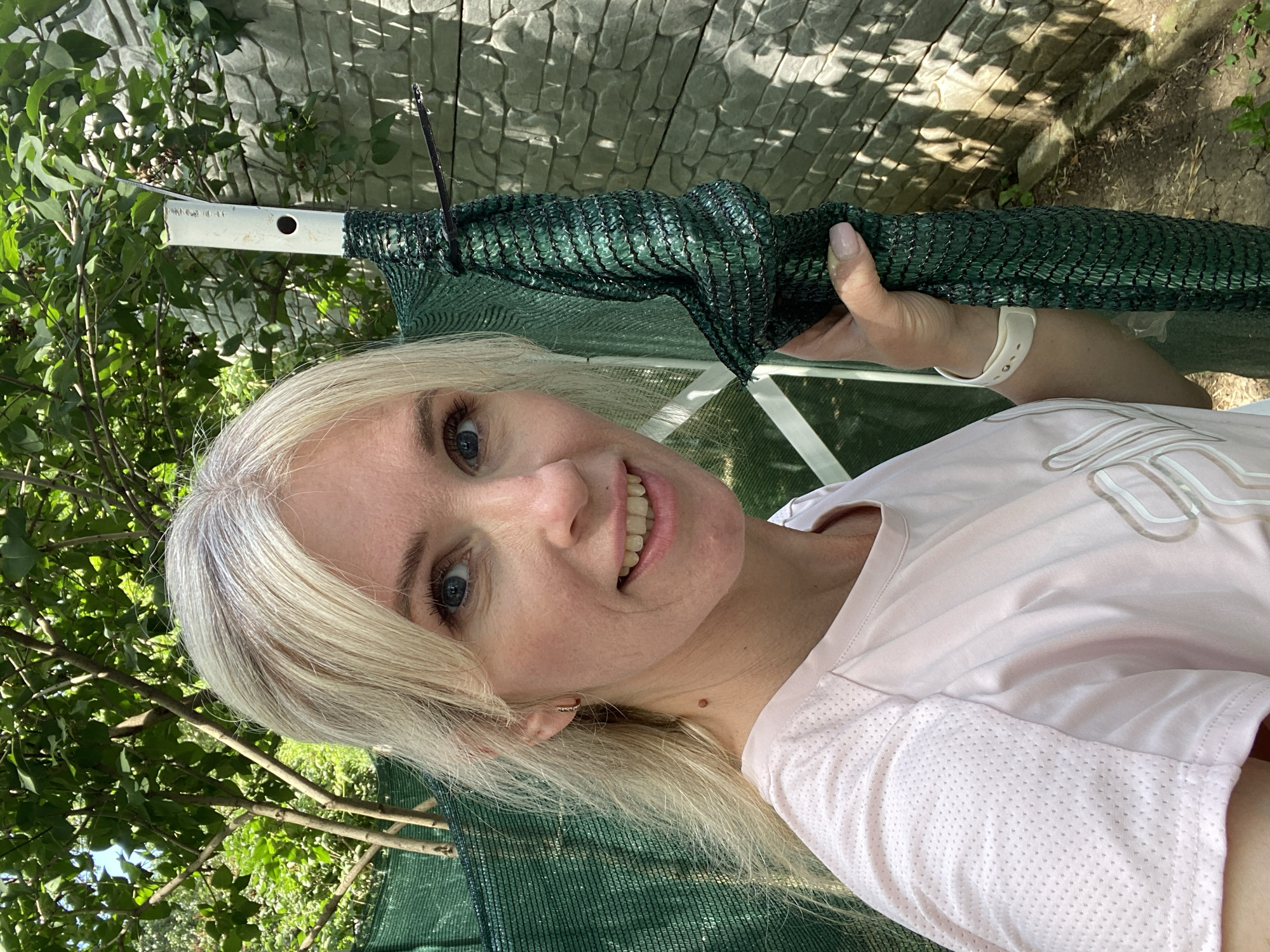 The media file is a portrait of a woman with blonde hair and blue eyes, taken outdoors in a backyard setting. She is smiling and looking at the camera. The woman is wearing a light pink t-shirt with a textured neckline. She has a white watch on her left wrist.
The woman is holding a rolled-up piece of green netting, which appears to be some kind of garden netting or shade cloth. A white pole is also visible, partially obscured by the netting. In the background, there is a stone wall, lush green foliage from trees and bushes, and some dry ground. The lighting suggests it is daytime, possibly sunny, with shadows cast by sunlight filtering through the leaves. The overall mood is casual and relaxed. The image is oriented sideways, with the woman's head towards the left of the frame. There is no visible text in the image.
